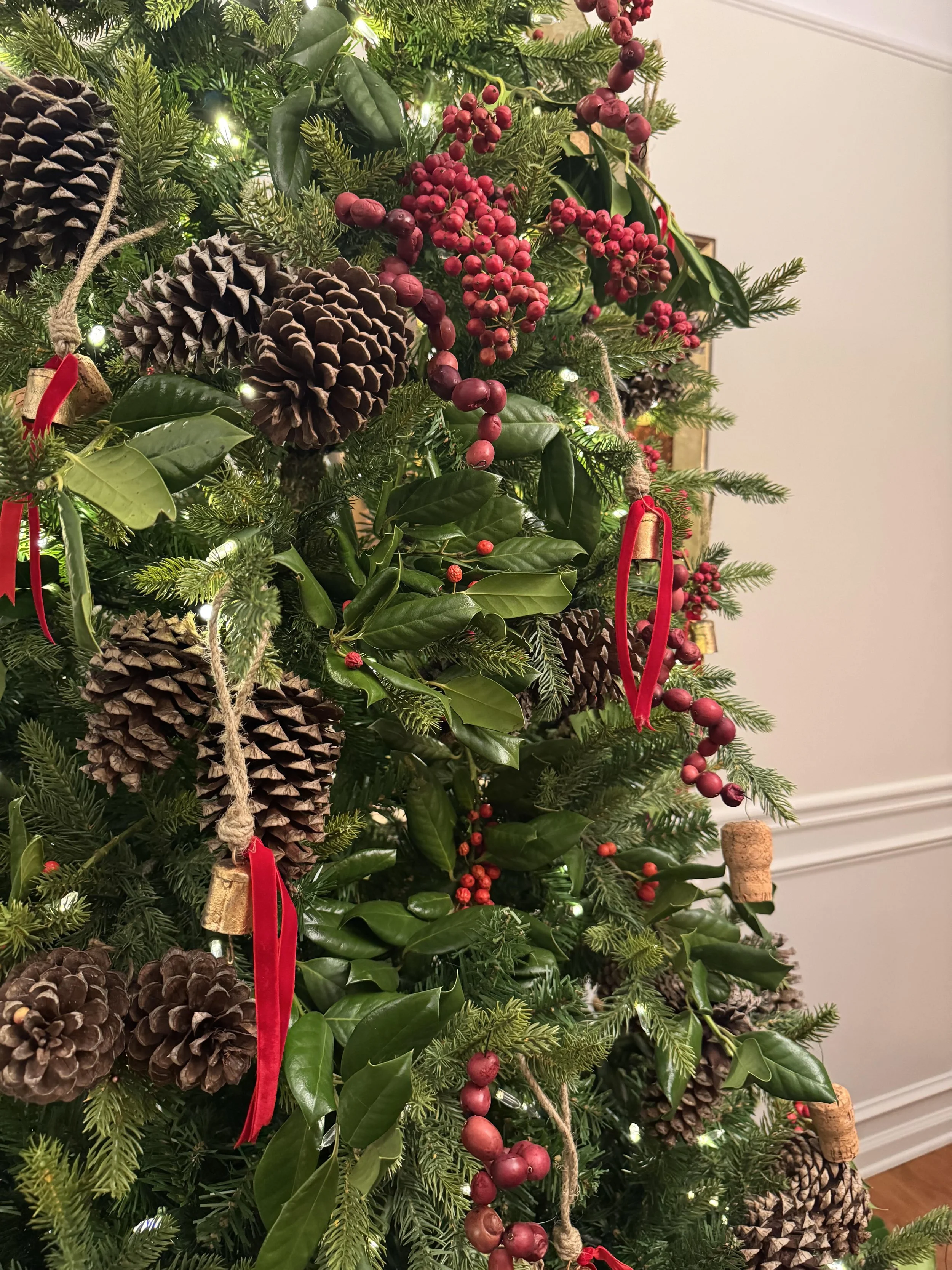 Close-up of a decorated Christmas tree with pinecones, red berries, greenery, string lights, red ribbons, and bells.