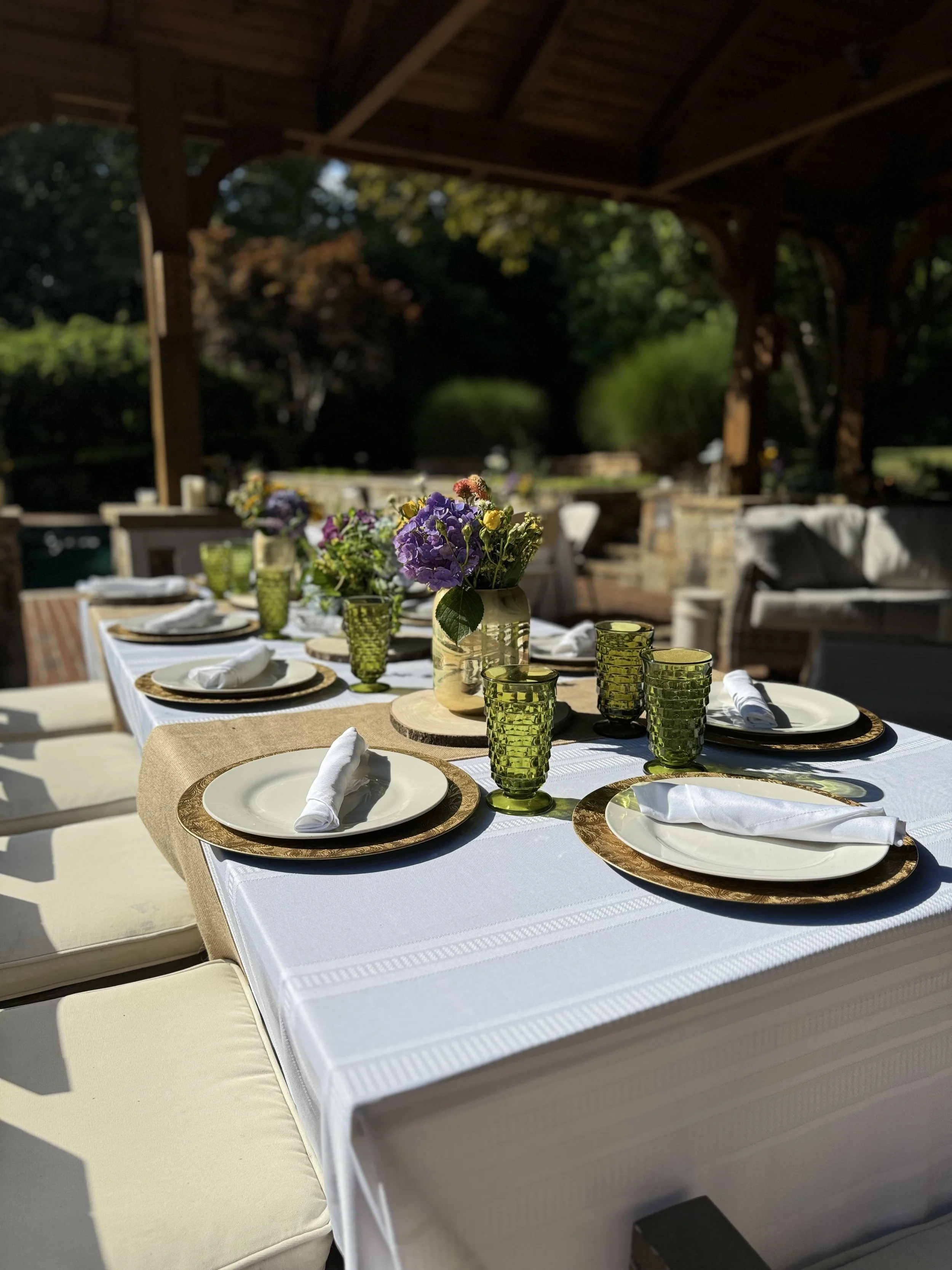 Elegant outdoor dining table with white linens, gold-rimmed plates, green glassware, and purple flower centerpiece under a pergola