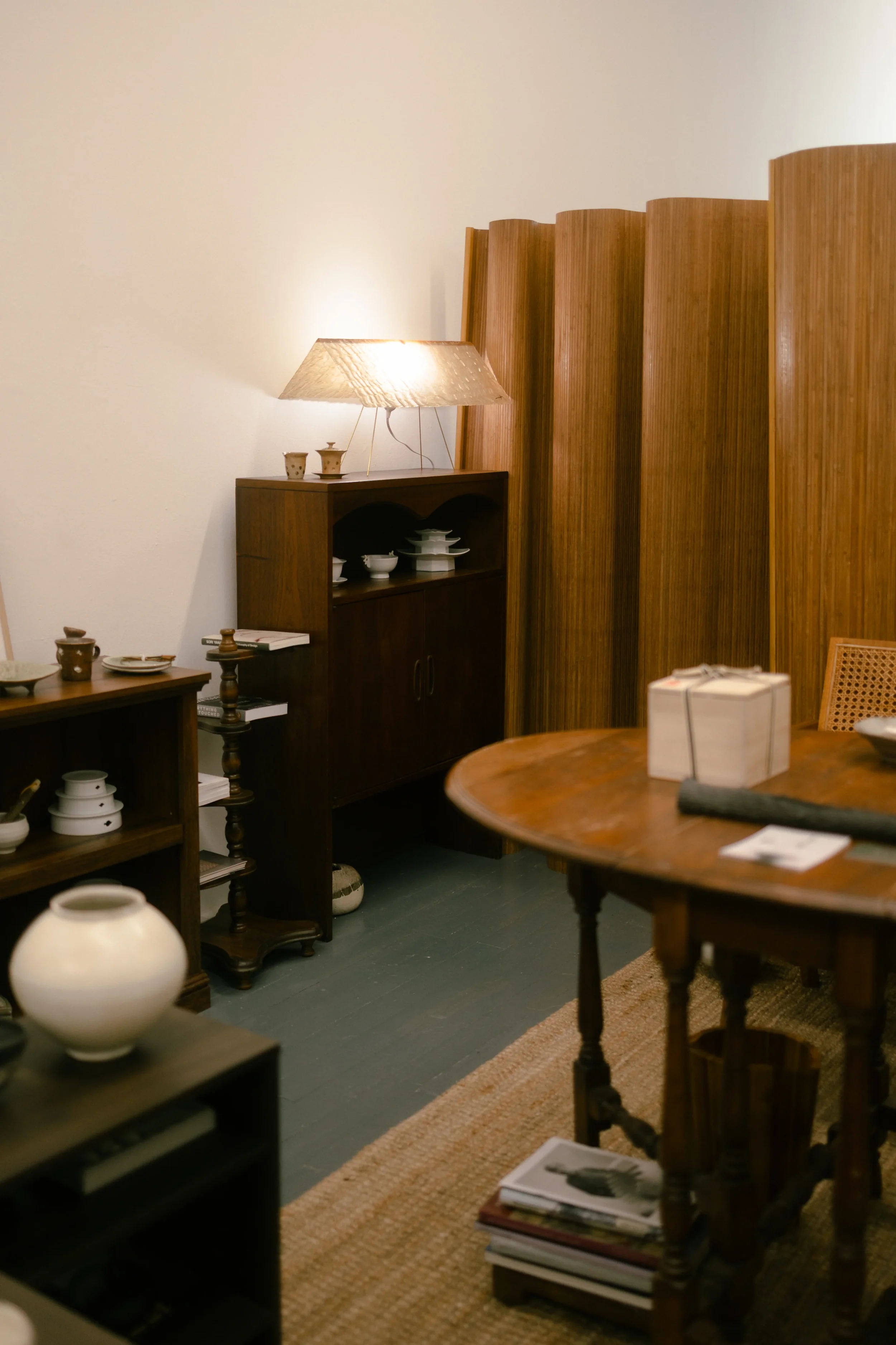 Interior of a room with wooden furniture, including a round table, bookshelves, and a cabinet. There are decorative items and a table lamp providing soft lighting.