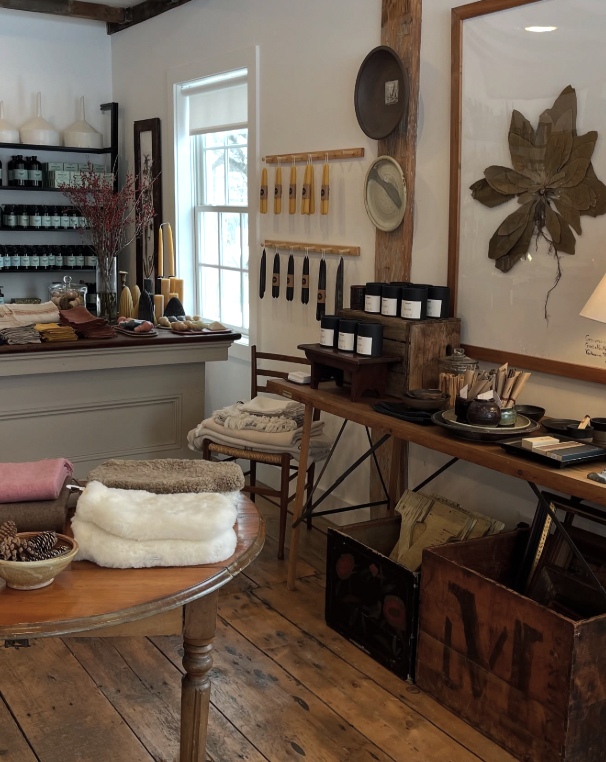 Interior of a cozy store with wooden flooring, displaying textiles, candles, and home decor items, with a large botanical print on the wall.