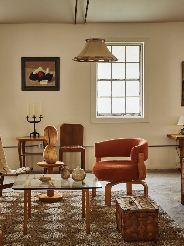 A cozy living room with a glass coffee table, a burnt orange armchair, a wooden mid-century chair, a small wooden side table with a candelabra, a framed abstract artwork on the wall, and a large window allowing natural light. A woven basket sits near the coffee table, and a vintage hanging ceiling lamp is overhead.