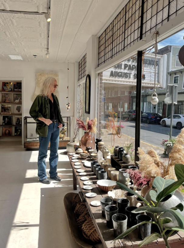 Woman browsing handcrafted pottery and vases inside a bright retail store with large front windows showing street view.