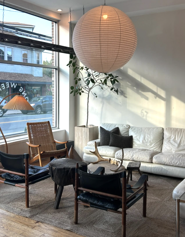 Living room with large window, white sofa, black pillows, a rattan armchair, a black lounge chair, a small table with decorative antlers, a hanging paper lantern, a potted plant, and sunlight casting shadows.