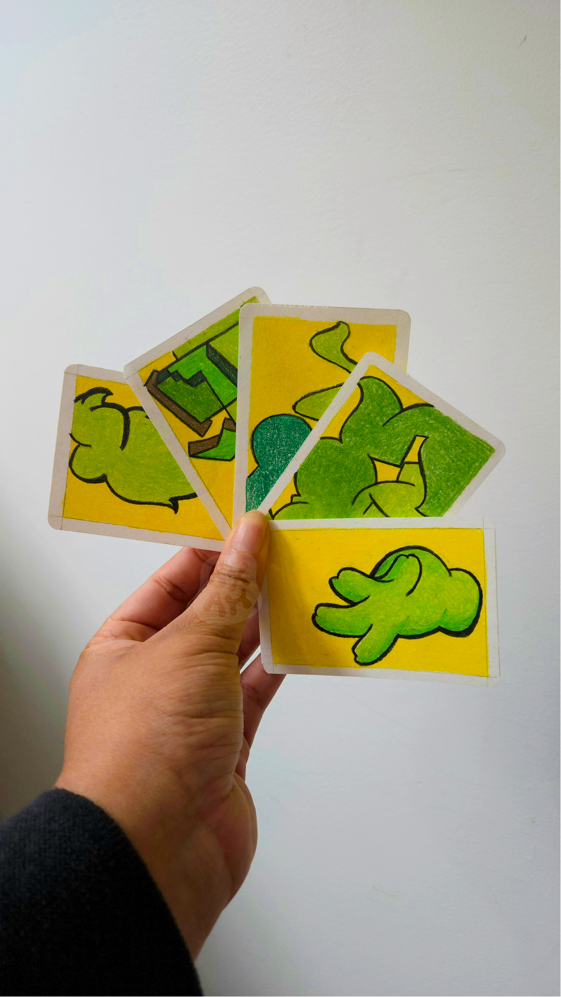 Hand holding colorful illustrated cards featuring green animals and landscapes against a plain white background.
