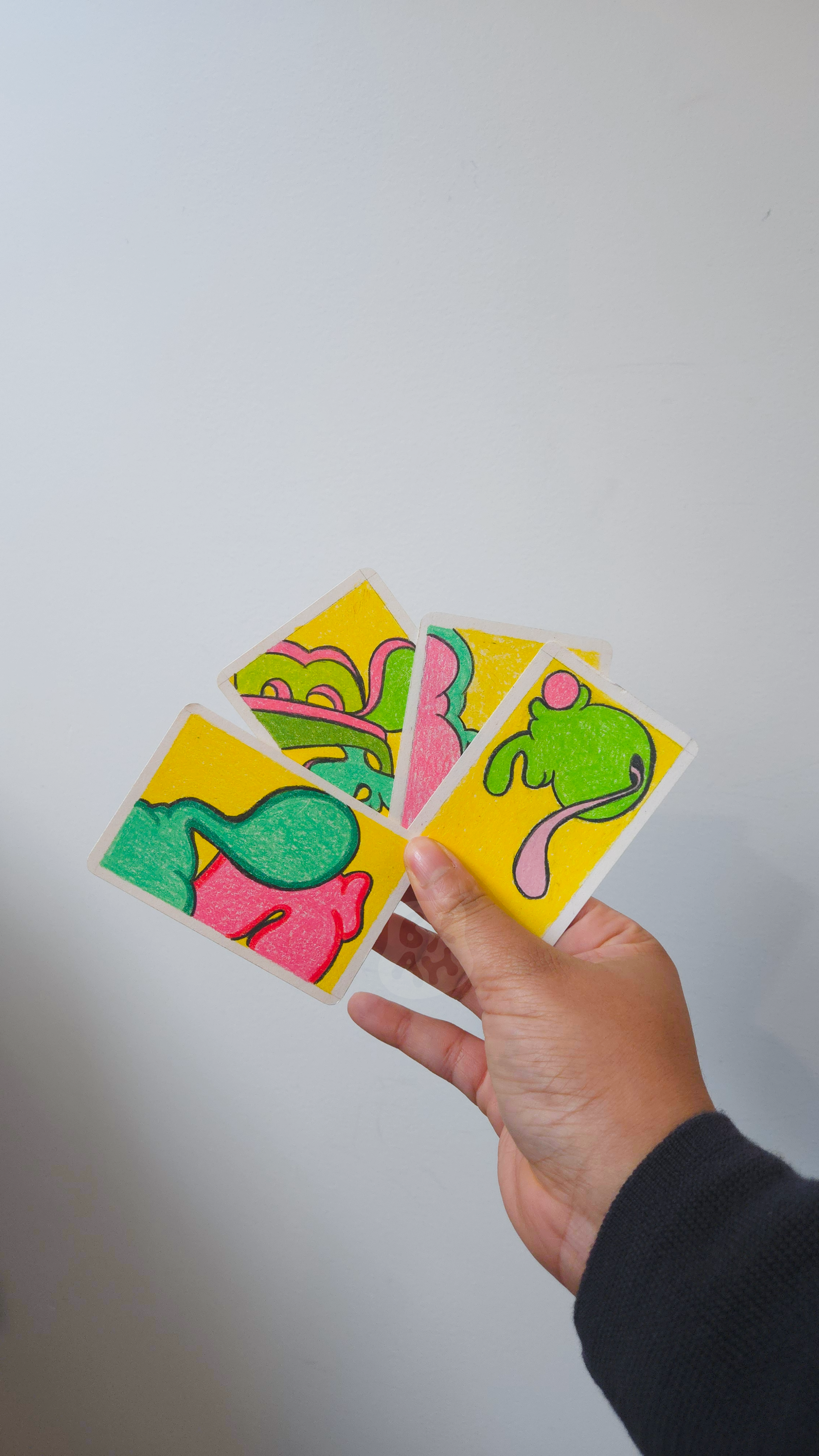 A hand holding four colorful cards with abstract, cartoon-like illustrations of creatures on a plain gray background.