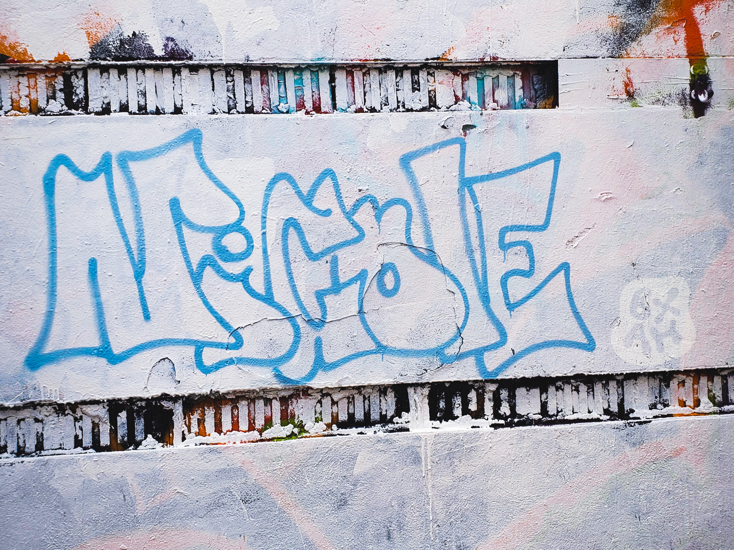 Graffiti tag in blue spray paint on a white wall with small vents or slats above and below the graffiti.