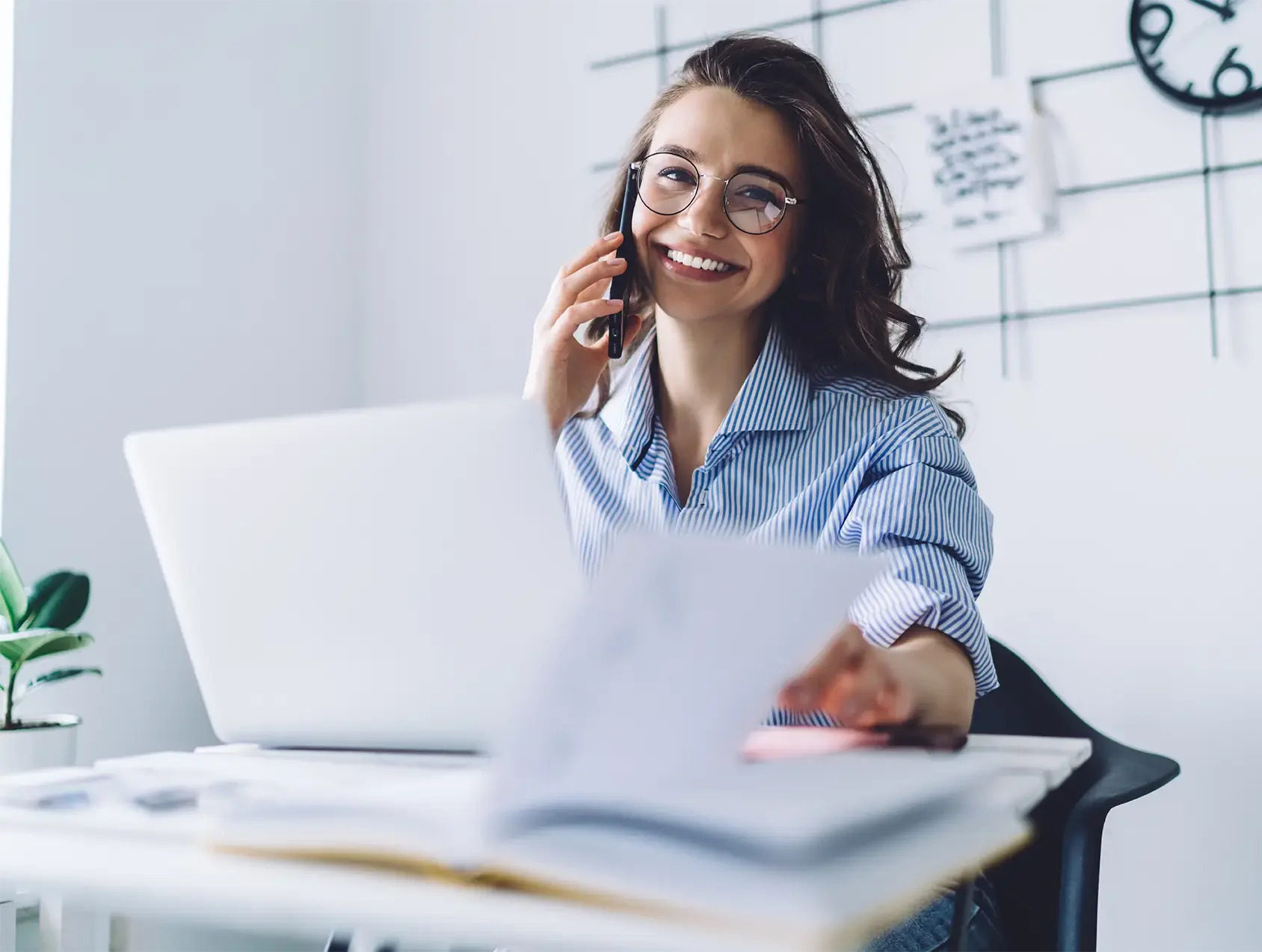 A young executive woman with glasses, smiling, sitting at a desk in an office, talking on a mobile phone, and holding a piece of paper.