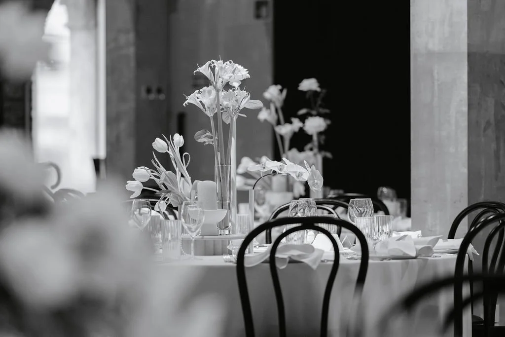 Elegant dining table with white flowers in tall vases, set with glassware, white napkins, and plates at a formal event in a modern interior.