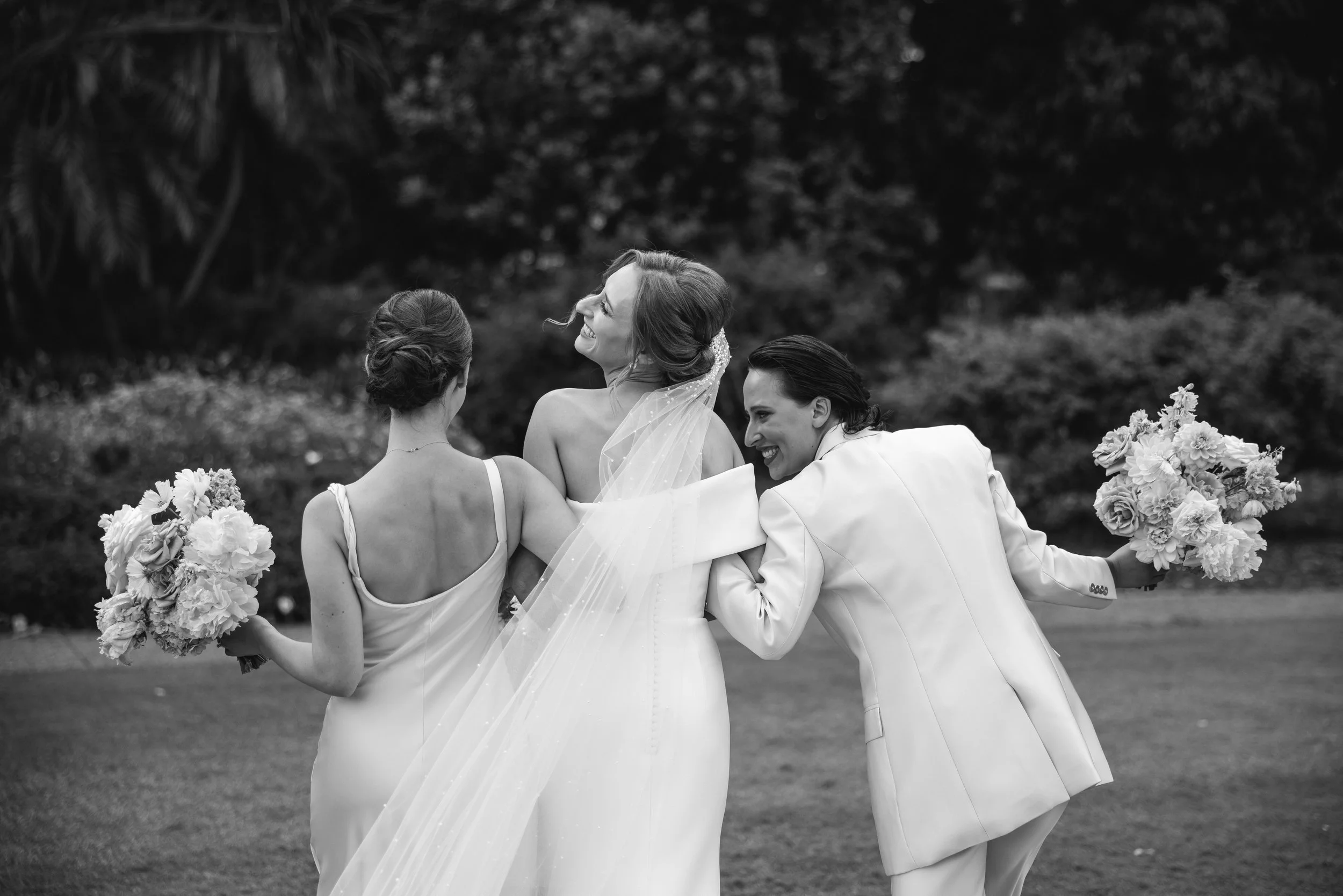 Black and white photo of a bride with two women, one holding a bouquet of flowers, all smiling and laughing outdoors.