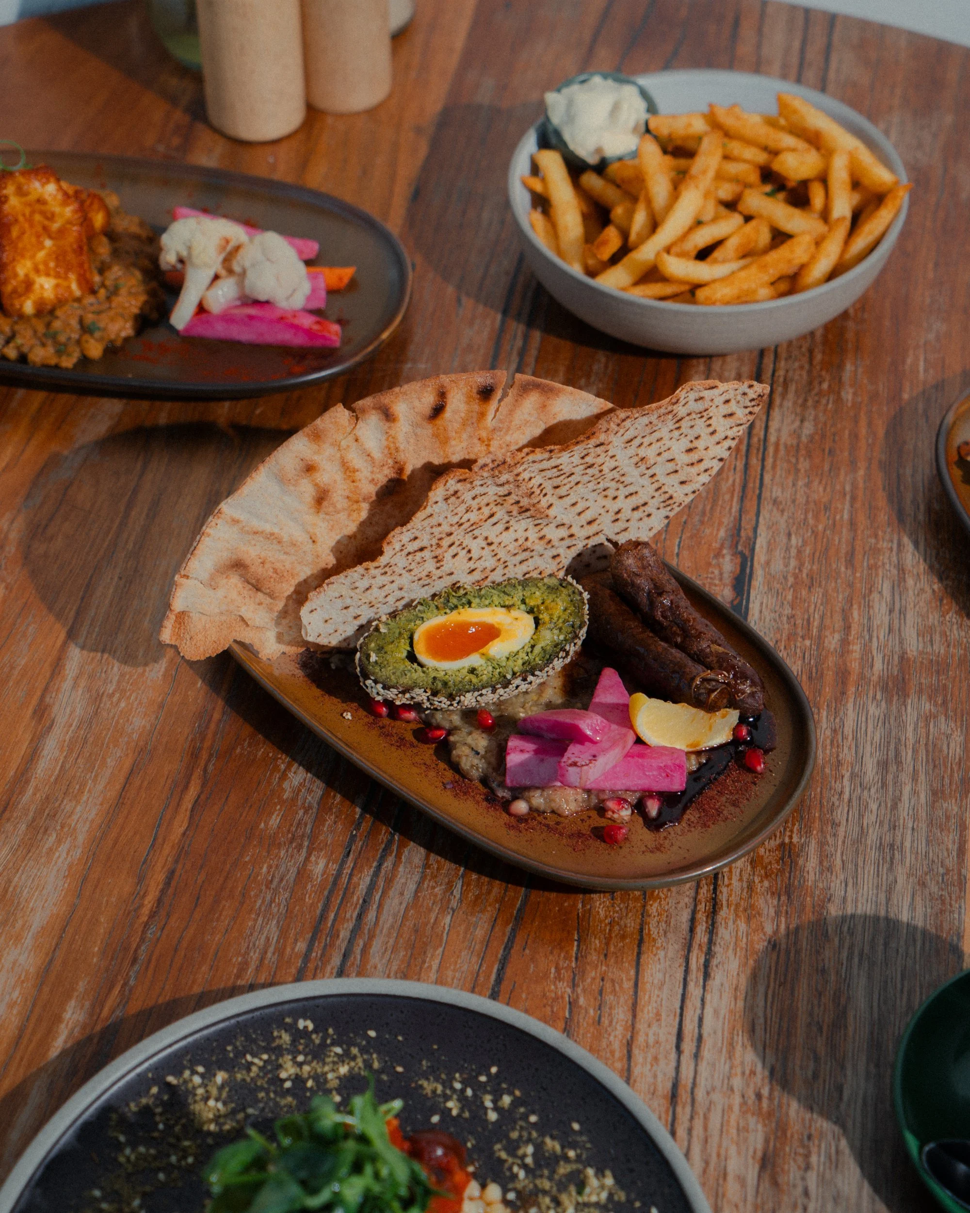 A plate with a Scotch egg, sausage, pink pickled vegetables, lemon wedge, and pita bread on a wooden table, with a bowl of French fries and other dishes in the background.