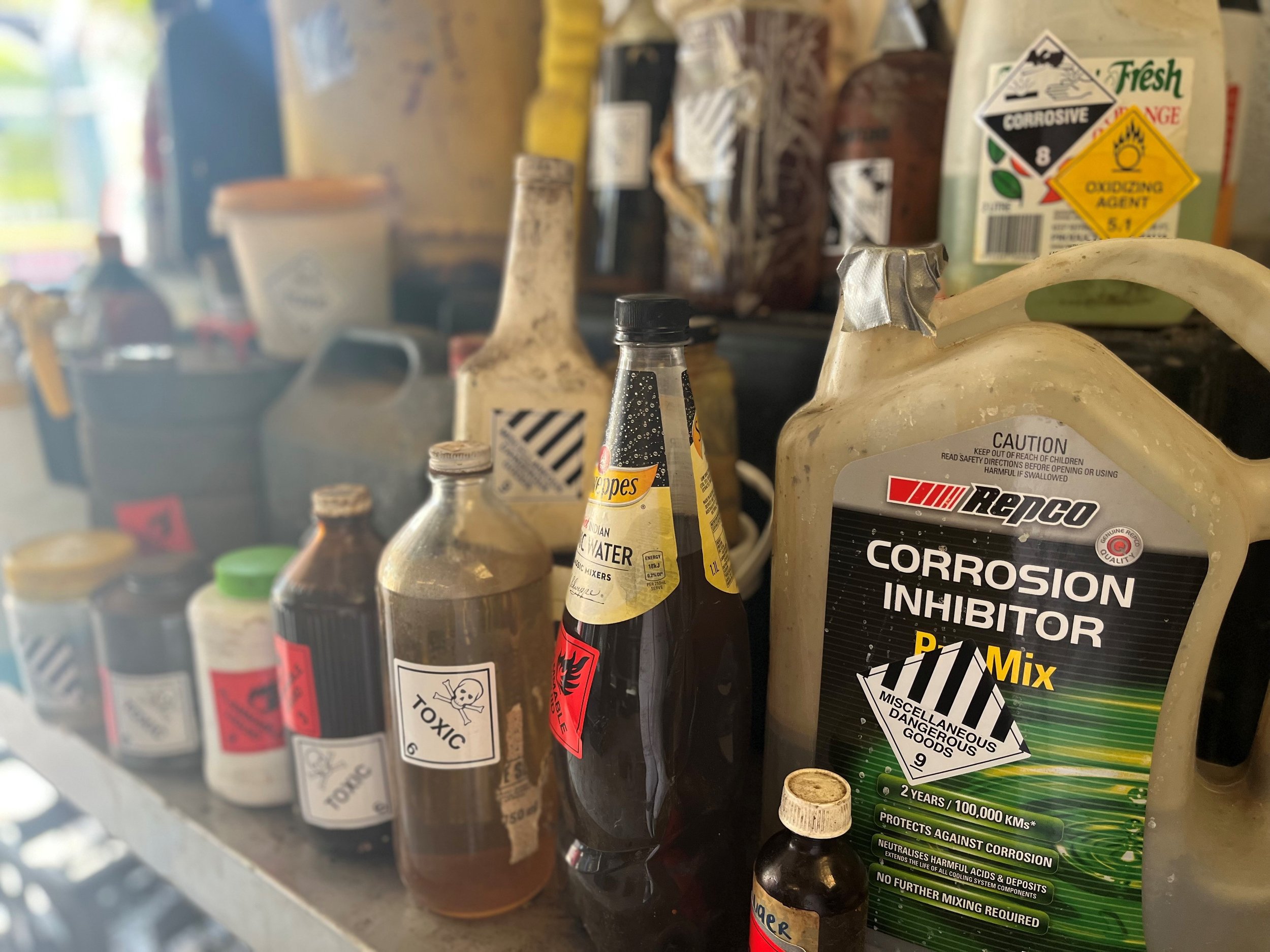 Collection of chemical containers, including bottles labeled with warnings like "toxic" and "corrosive." Prominent "Corrosion Inhibitor" container in front.