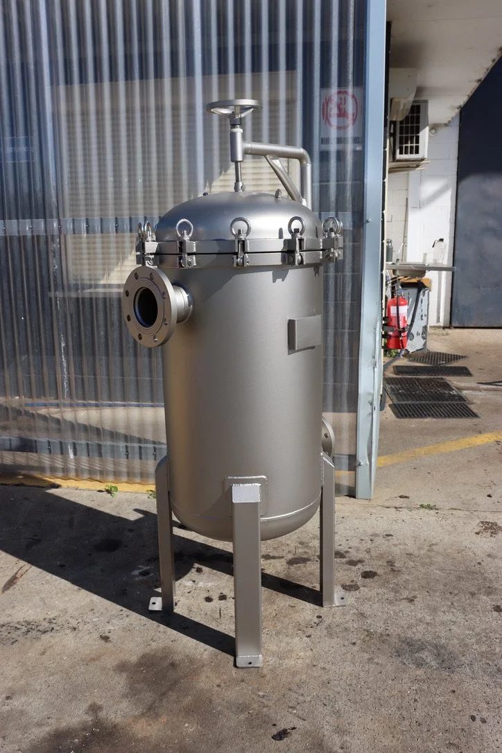 Industrial stainless steel filter or pressure vessel with a top handle and a side flange, standing outdoors on concrete near a metal building.
