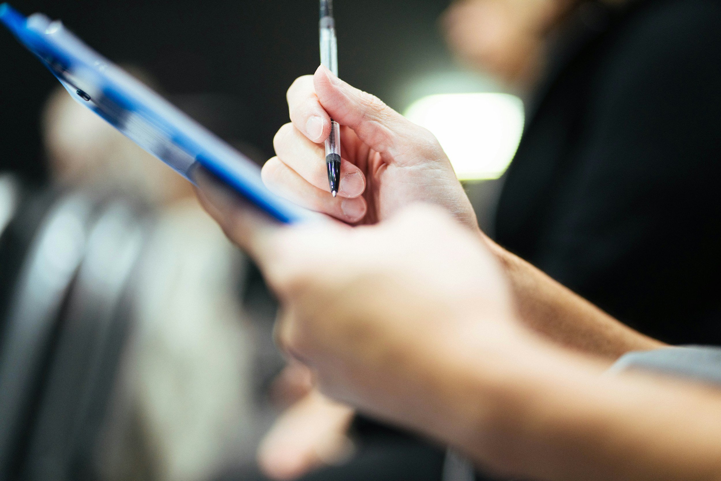 Close-up of a person's hand holding a black ink pen and writing on a surface, with a blurred background.