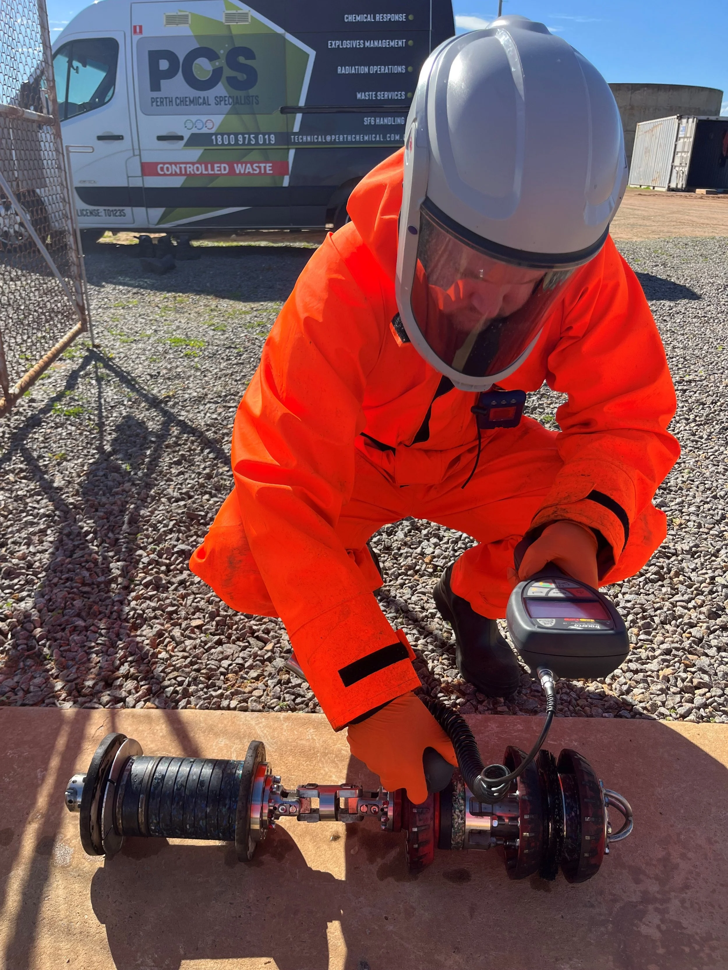 Person in orange protective suit inspecting equipment outdoors, next to a van with company branding and control waste signage.