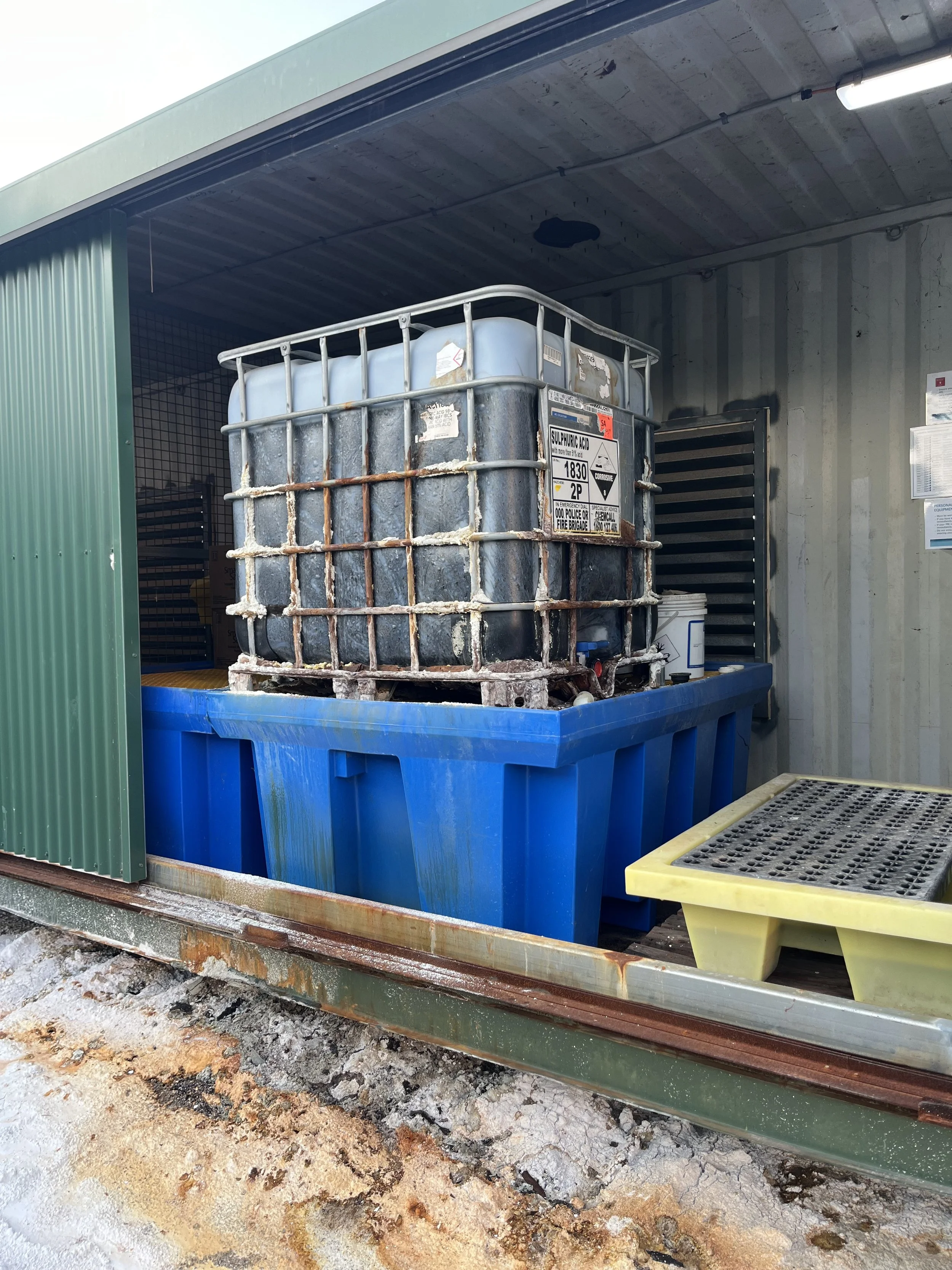 Large container labeled sulfuric acid inside a storage unit, placed on a blue spill containment pallet with a yellow grid pallet beside it.