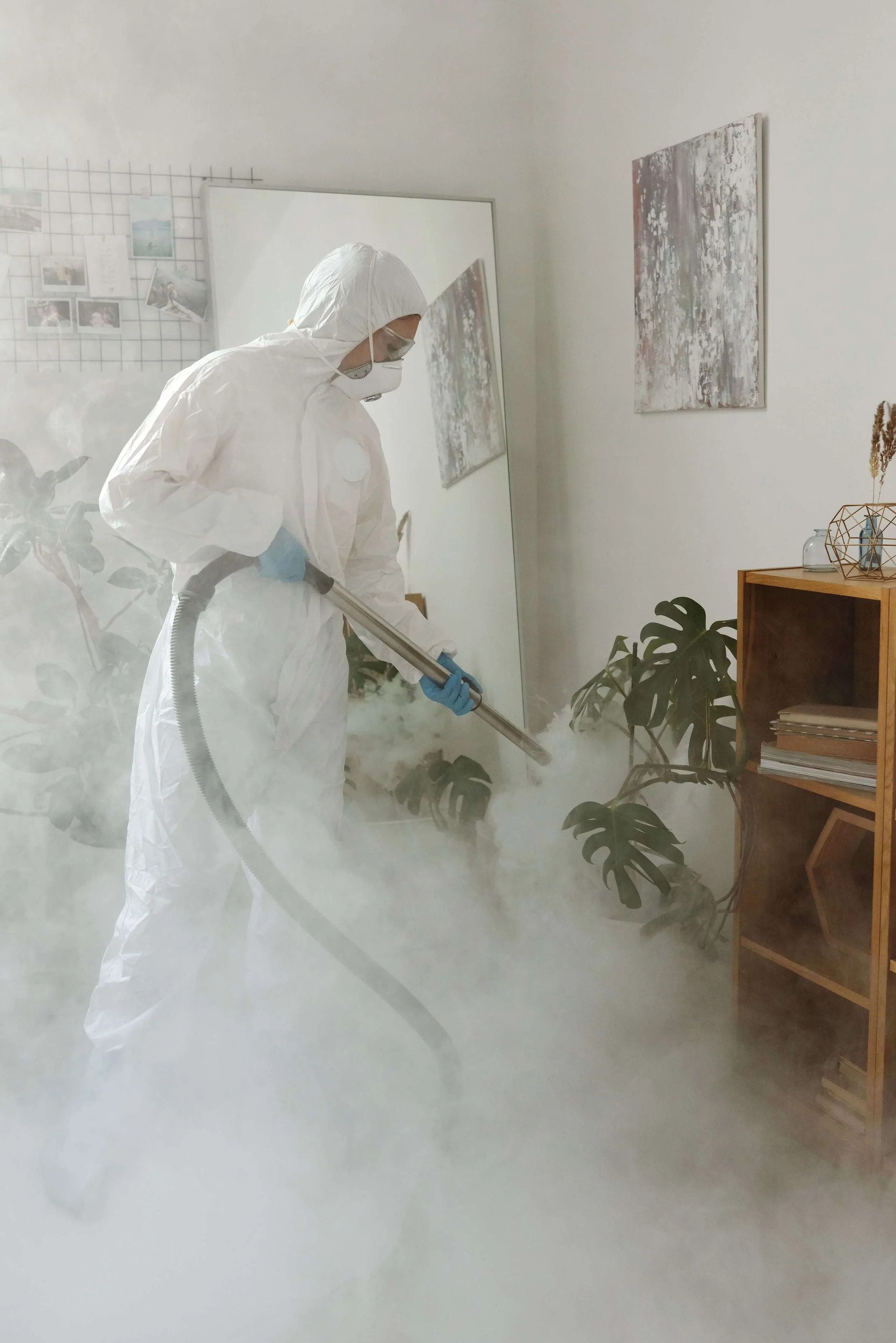 Person in protective suit, mask, and gloves using fogging equipment in a room with plants and modern decor.
