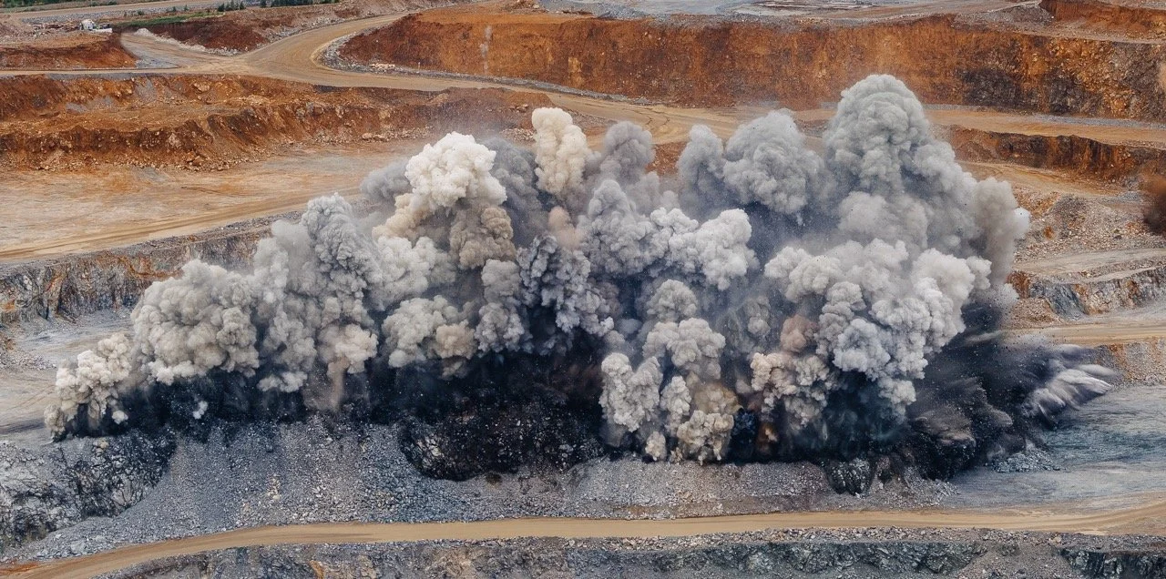 An open-pit coal mine explosion with clouds of black and gray dust and smoke erupting from the ground.