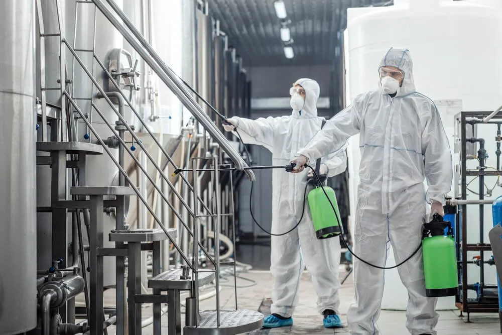 Two workers in protective suits and masks disinfect a large industrial facility using spray tools connected to green tanks.