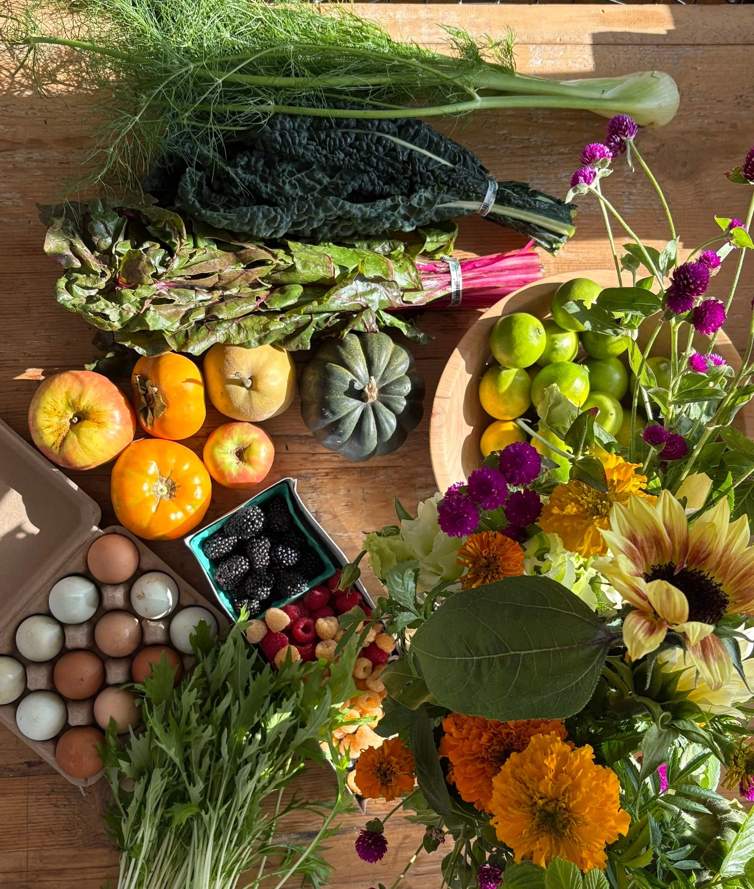 A variety of fresh vegetables, fruits, eggs, berries, and flowers arranged on a wooden table, including apples, tomatoes, acorn squash, leafy greens, and colorful blooms.