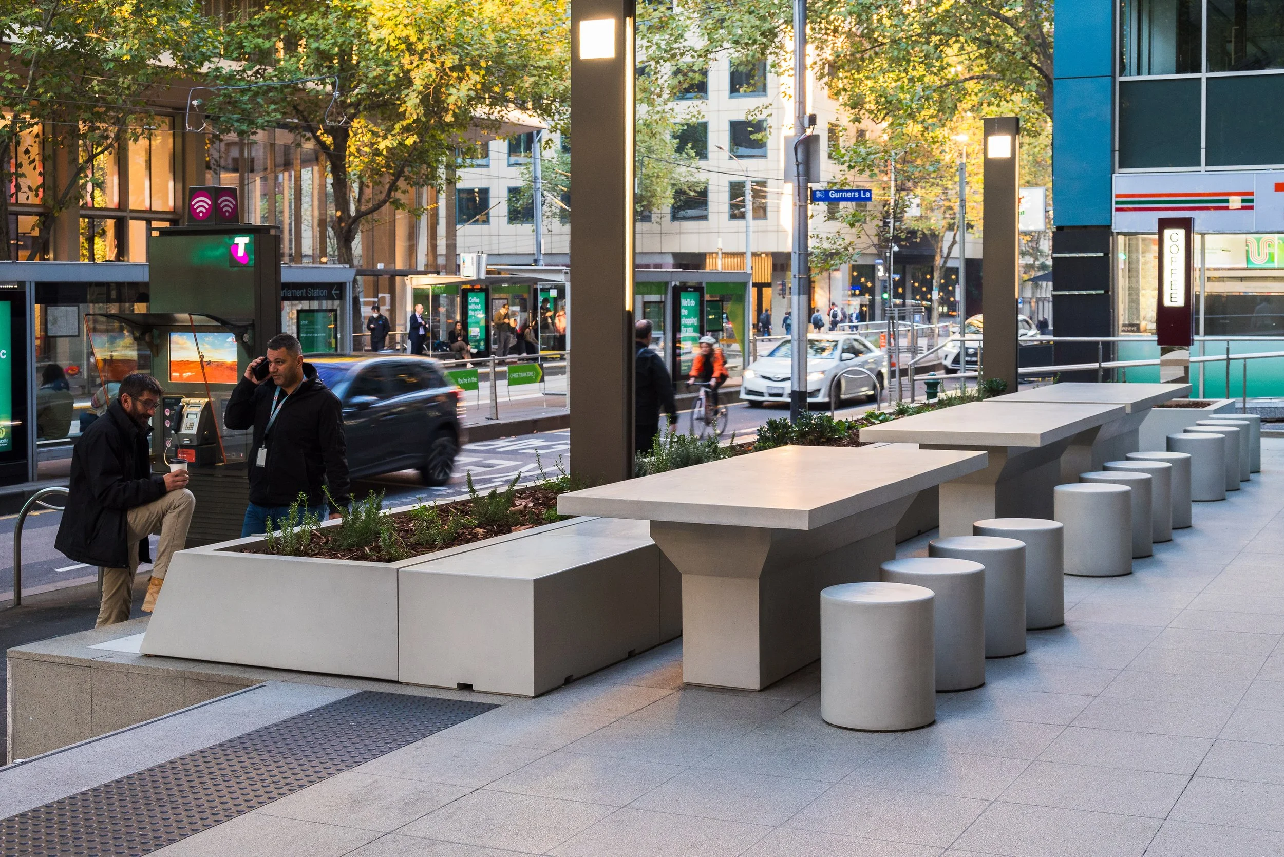 Urban streetscape with custom GRC street furniture including cylindrical stools, a robust outdoor table, a linear backless bench, and a low trapezoidal GRC planter alongside the pedestrian walkway.