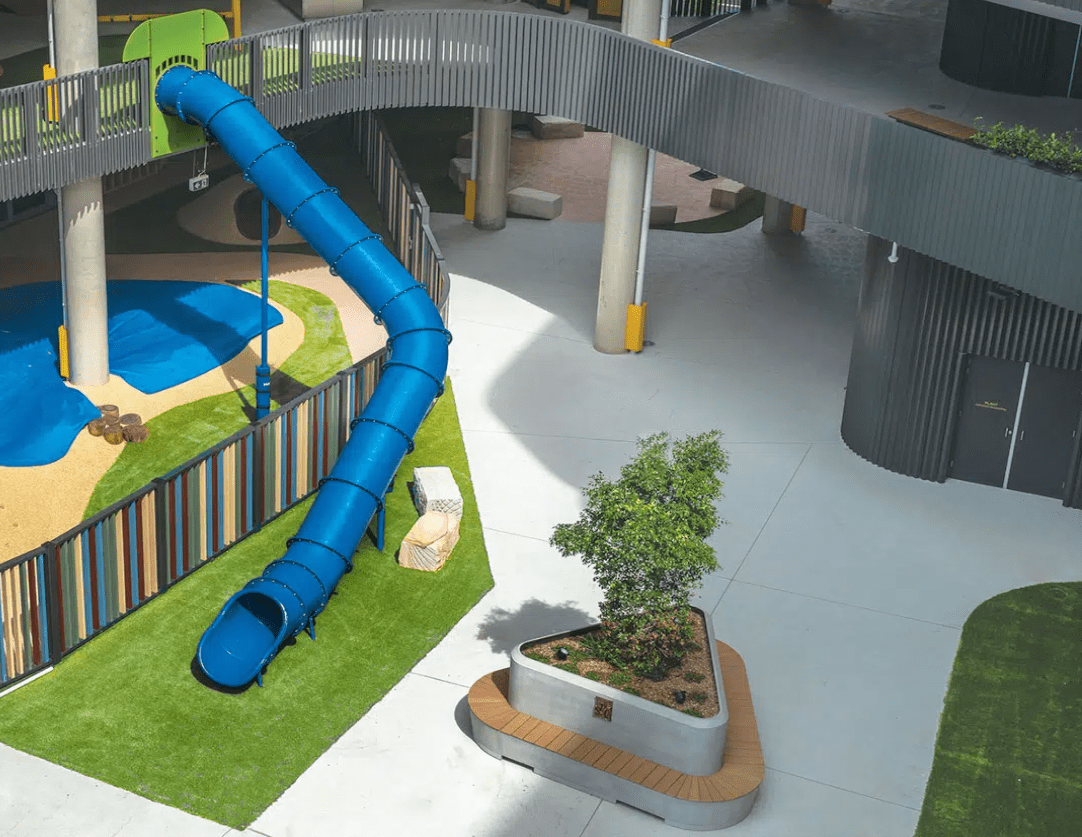 Ground-level outdoor space featuring custom round GRC planter benches surrounded by playful landscaping, colourful paving, and communal gathering areas.