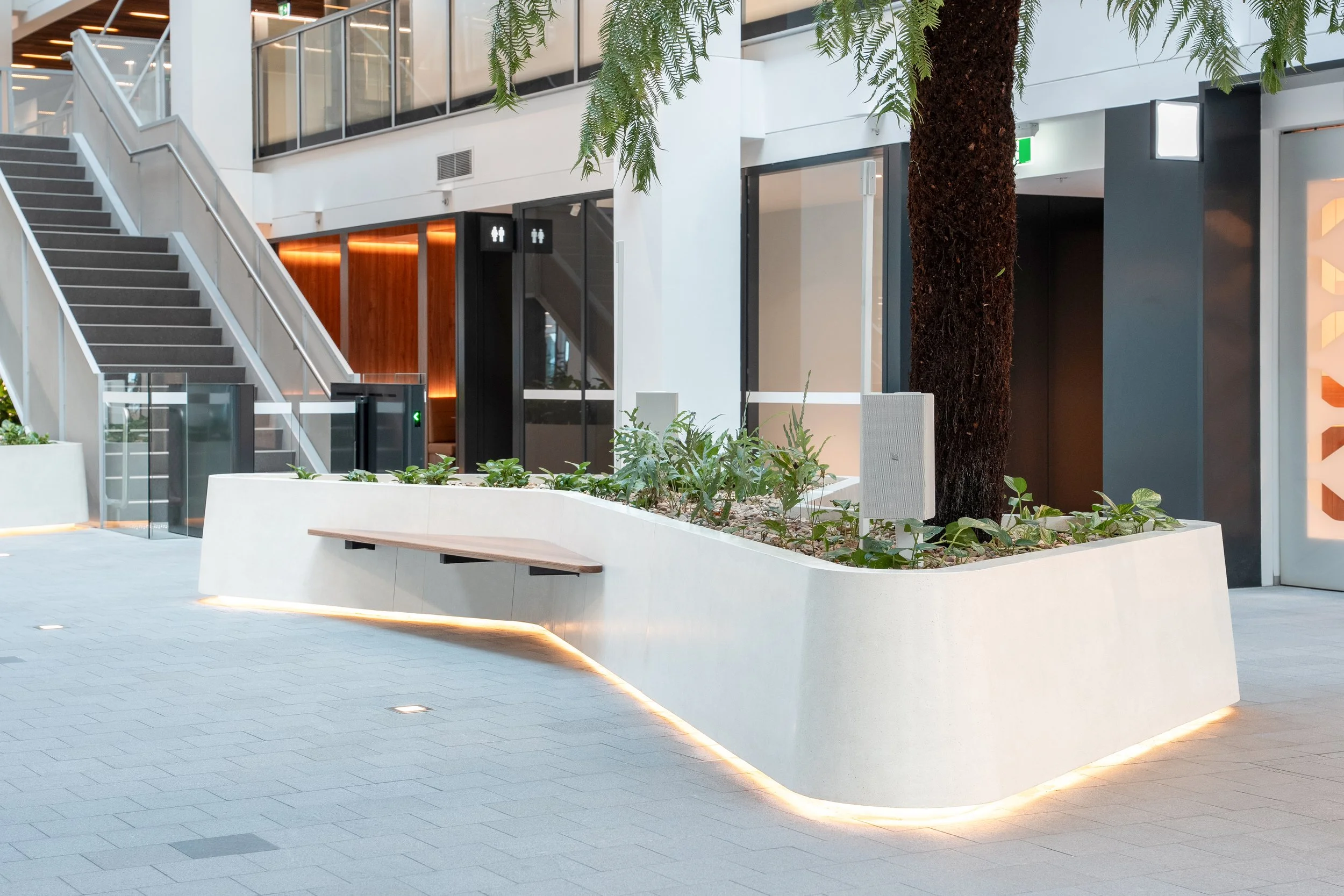 Indoor shopping mall or office space with modern white planter containing green plants and a large tree, with stairs, glass barriers, and shop entrances in the background.