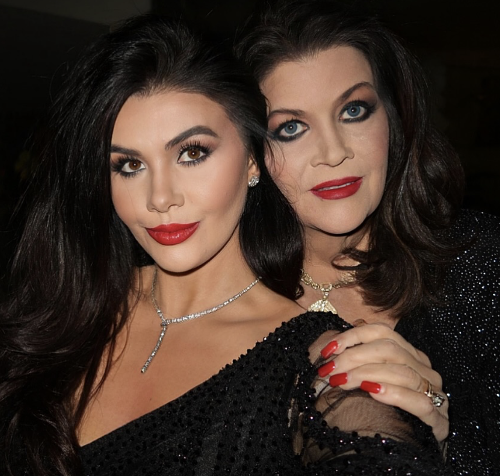 Two women with dark hair and bright makeup, dressed in black, posing closely together for a photo, with one woman holding her hand near her chest.