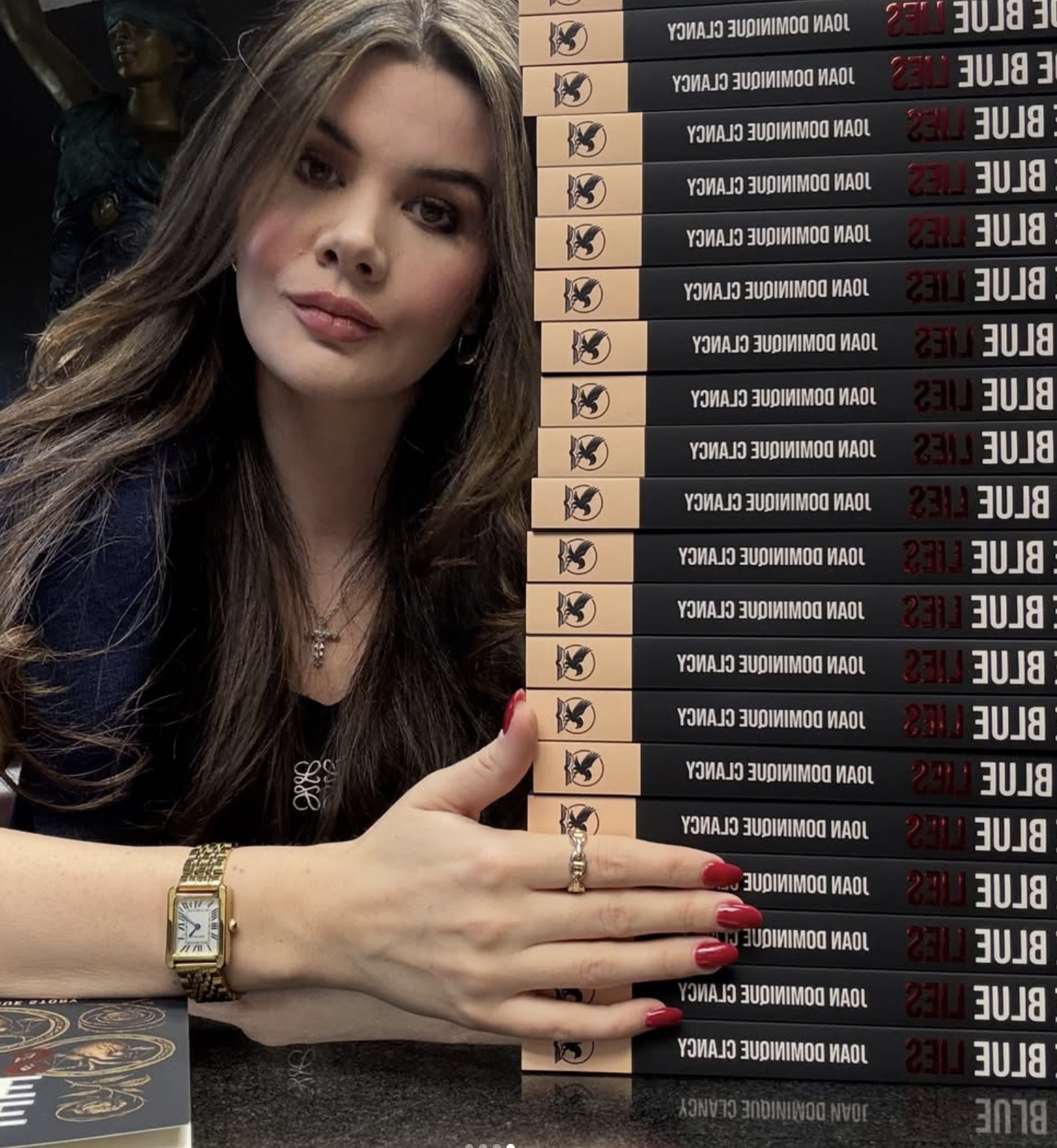 A woman with long brown hair, wearing a gold watch, a ring, and jewelry, is sitting at a table with her hand resting on the table, showing her red painted nails. She is next to a stack of books titled 'Blue Lies' by Joan Domi­nique Clancy, with the b