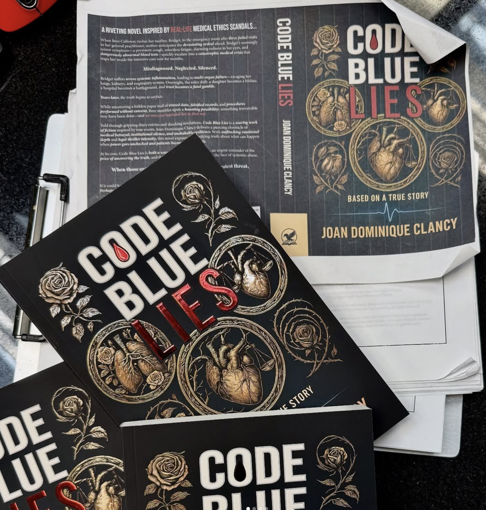 Multiple copies of a book titled 'Code Blue Lies' with gold and red illustrations of a human heart and roses, placed on top of a pile of papers and documents.
