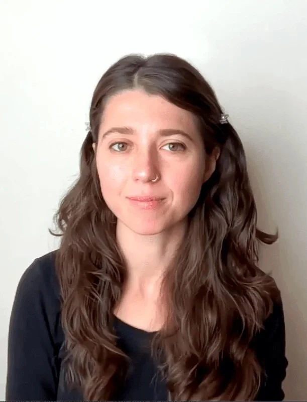 Photograph of Joy Deva Baglio, a young woman with long wavy brown hair and a nose ring, wearing a black top, standing in front of a plain white wall.