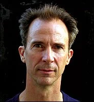 A portrait of a man with dark, slightly disheveled hair and a serious expression, against a black background.