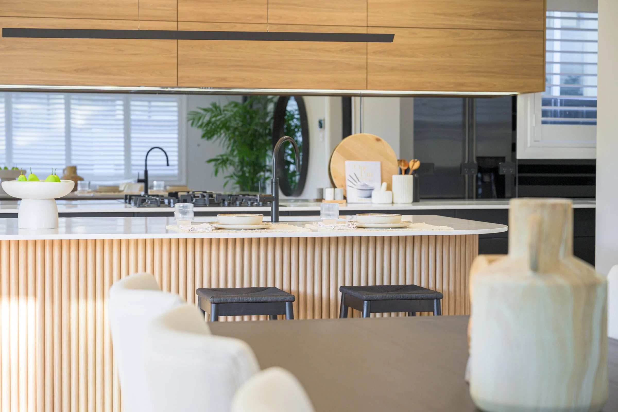 Modern kitchen with dining area, featuring a kitchen island with two black stools, a white vase with green apples, and a variety of kitchen utensils and decor, including wooden cutting boards and a plant in the background.