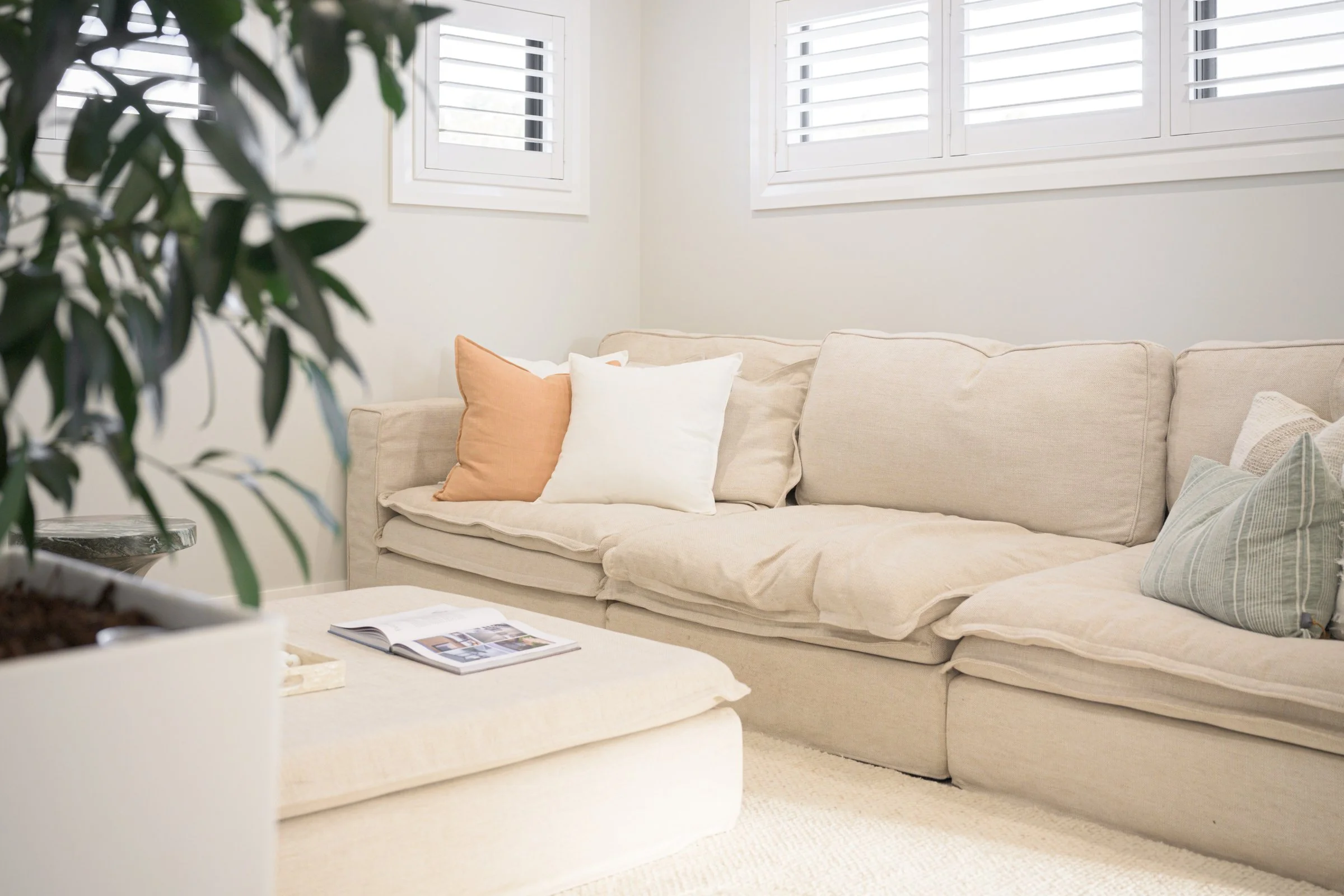 A beige couch with four pillows in cream, peach, green, and beige colors in a bright living room with white walls, windows, and plantation shutters.