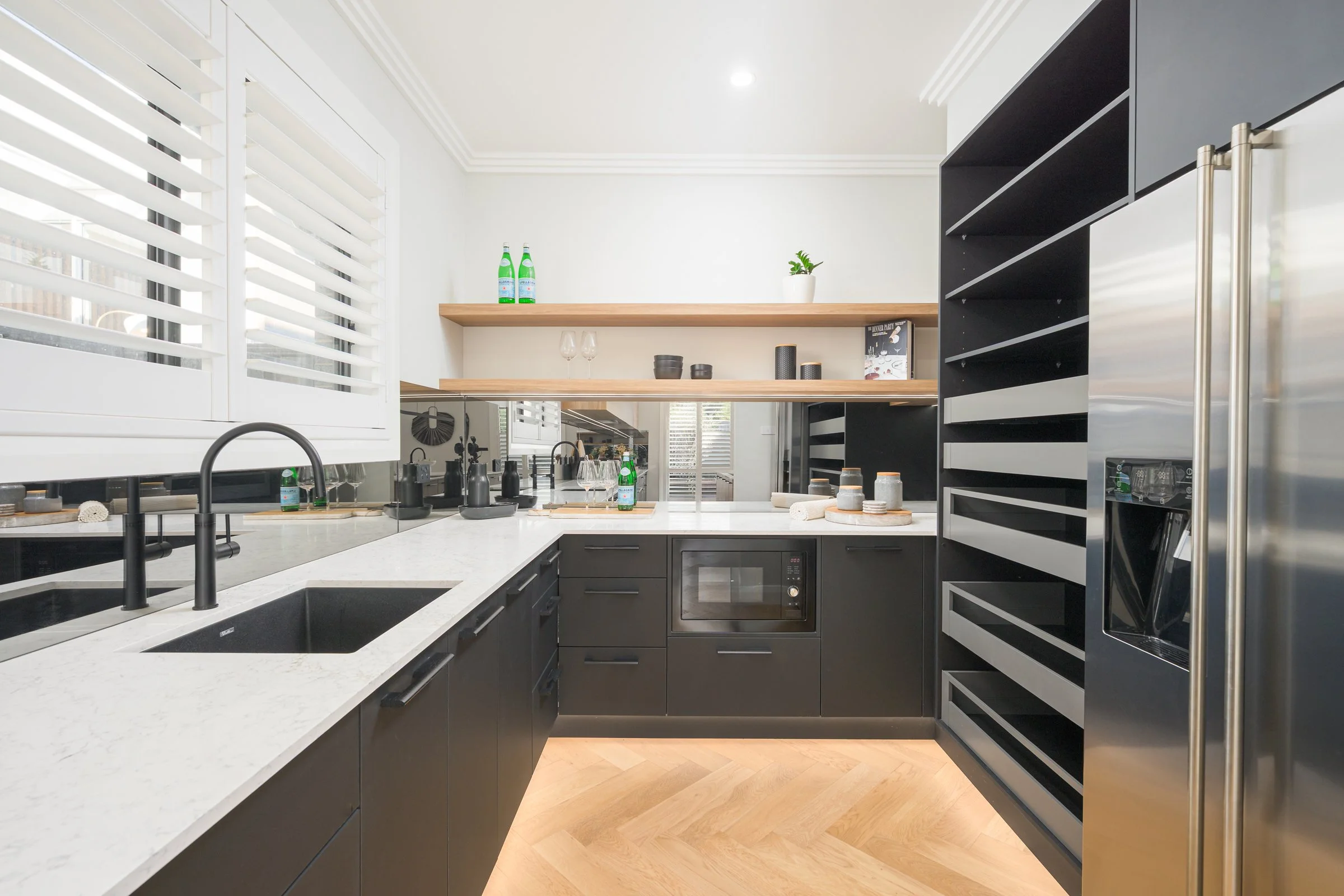 Modern kitchen with black cabinets, white countertops, and open wooden shelves. It features a stainless steel refrigerator, microwave, and black sink with a black faucet. Decor includes bottles, glasses, bowls, and potted plants, with natural light c