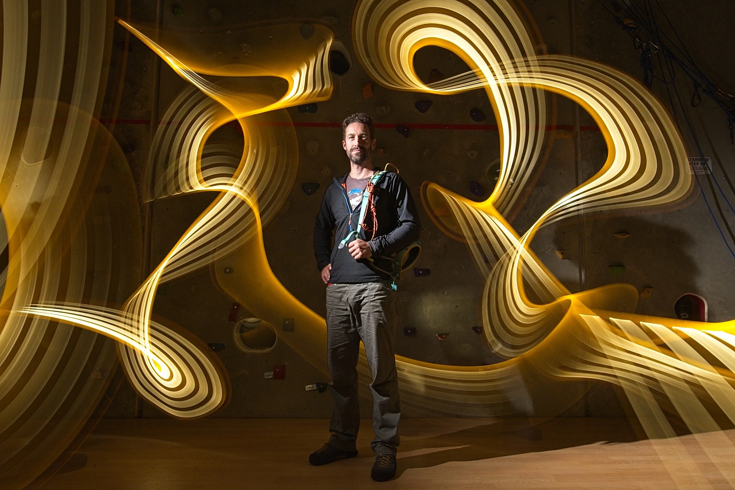 A man wearing outdoor gear stands in front of a rock climbing wall with abstract yellow light trails swirling around him.