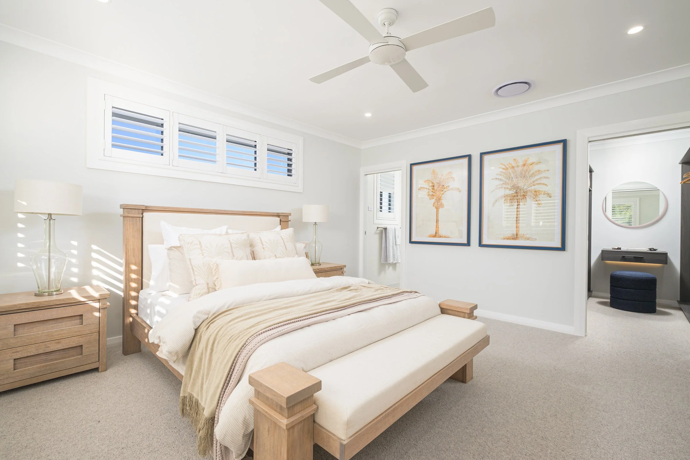 A bedroom with a large bed, wooden nightstands with lamps, wall art of palm trees, and a doorway leading to another room with a mirror and a stool.