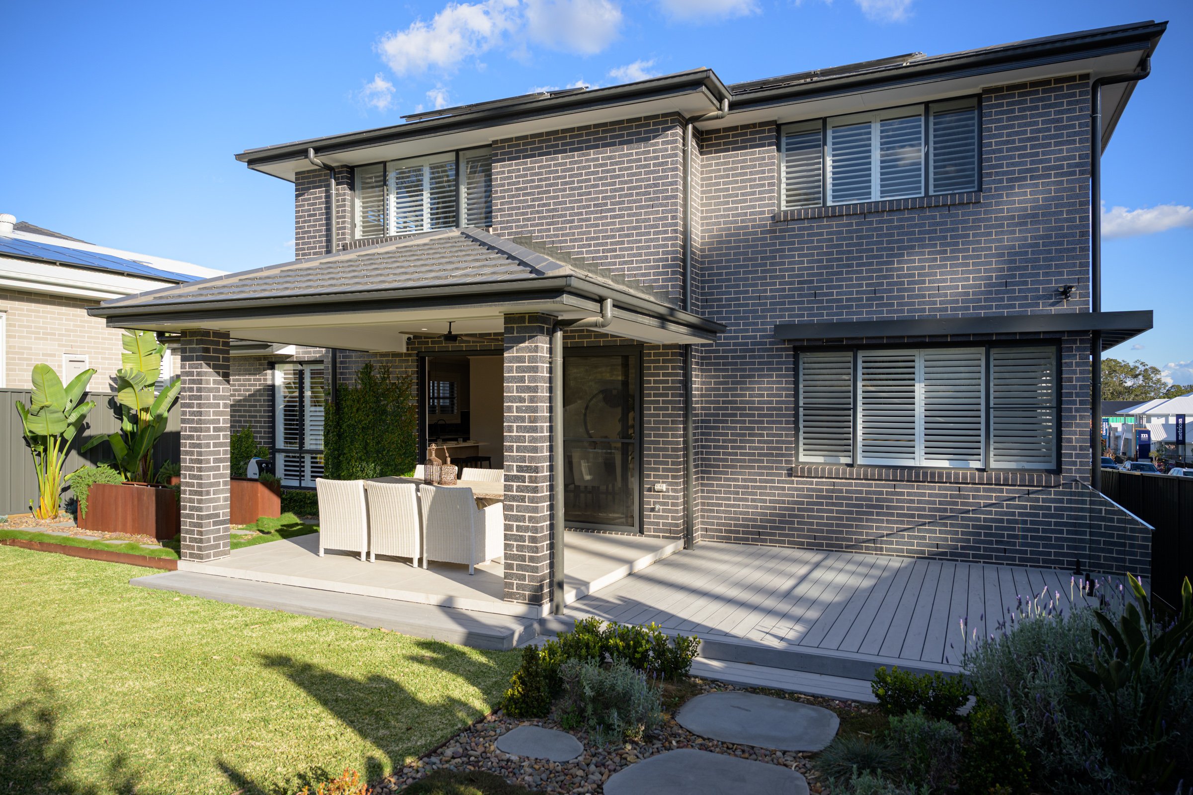 Modern two-story house with dark brick exterior, large windows with shutters, and a covered patio with outdoor furniture, surrounded by a landscaped yard with plants and a lawn.