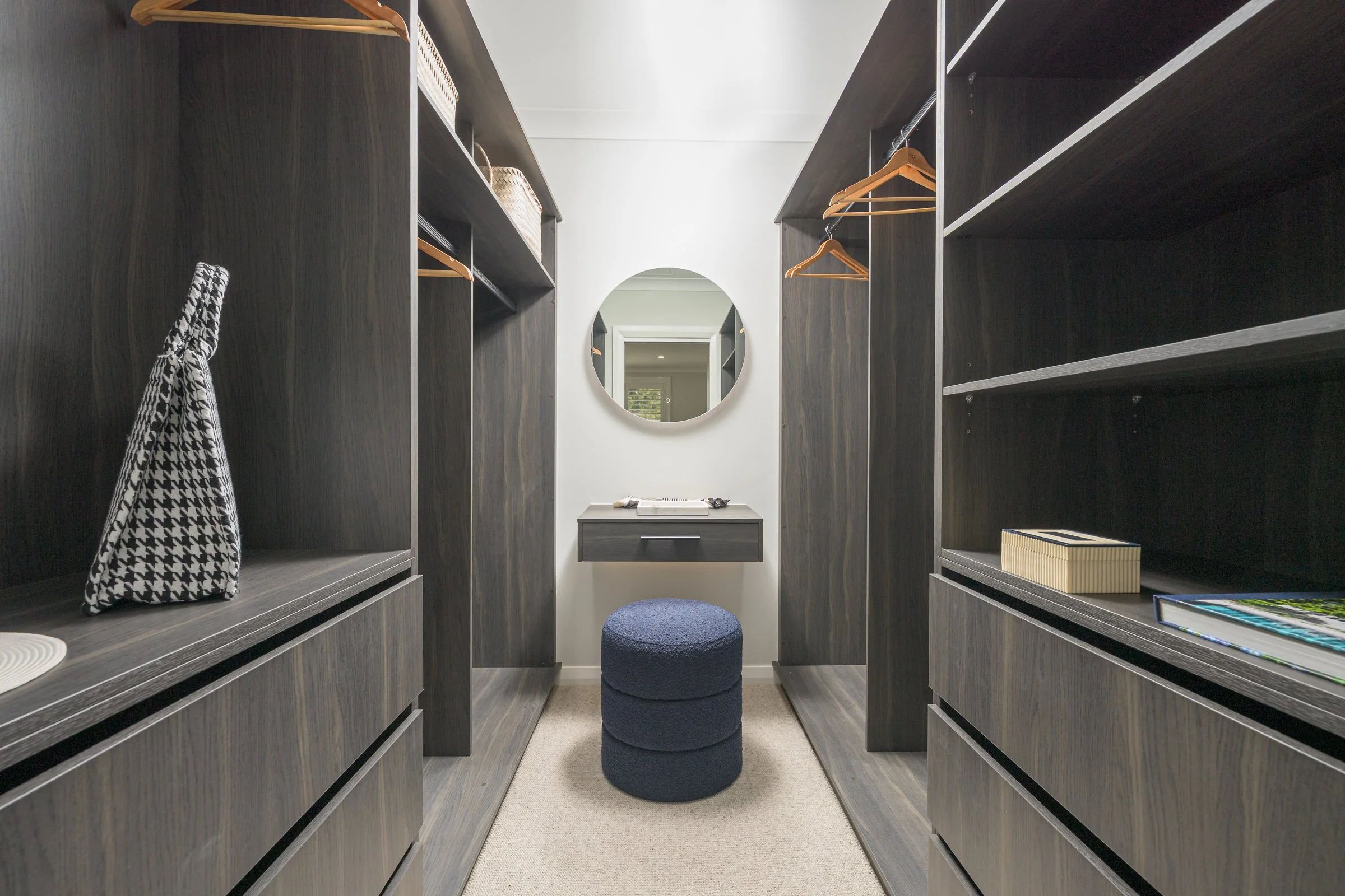 A walk-in closet with dark wood shelving and drawers on both sides, a small dark grey vanity with a round mirror above, a blue upholstered stool in the center, and various items like books, boxes, and a patterned bag displayed on the shelves.