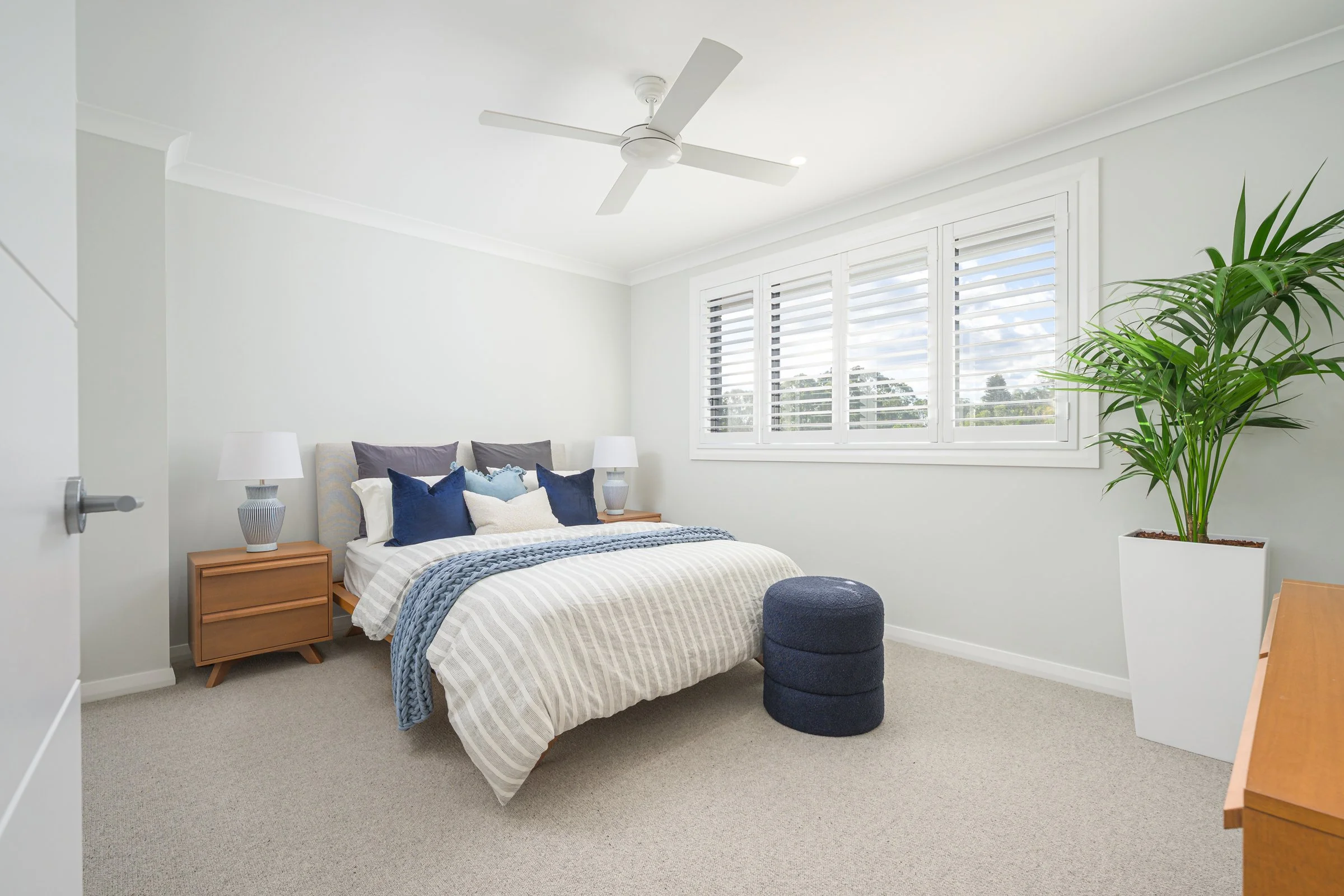 A bright bedroom with a bed featuring multiple pillows, wooden nightstands with lamps, a large window with white shutters, a large potted plant, and a small round ottoman.