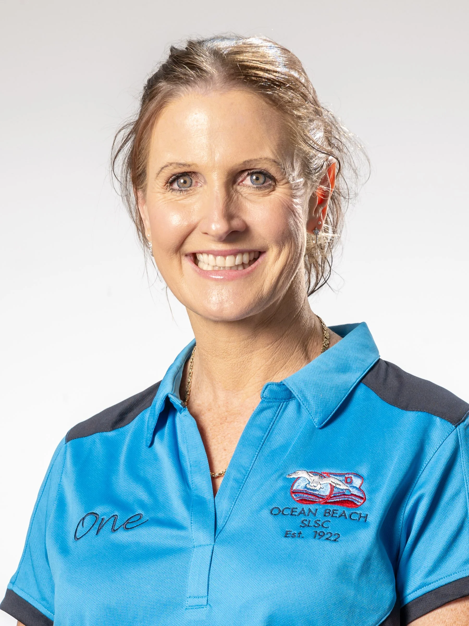 A smiling woman with short, light brown hair and blue eyes, wearing a blue polo shirt, posing against a plain white background.