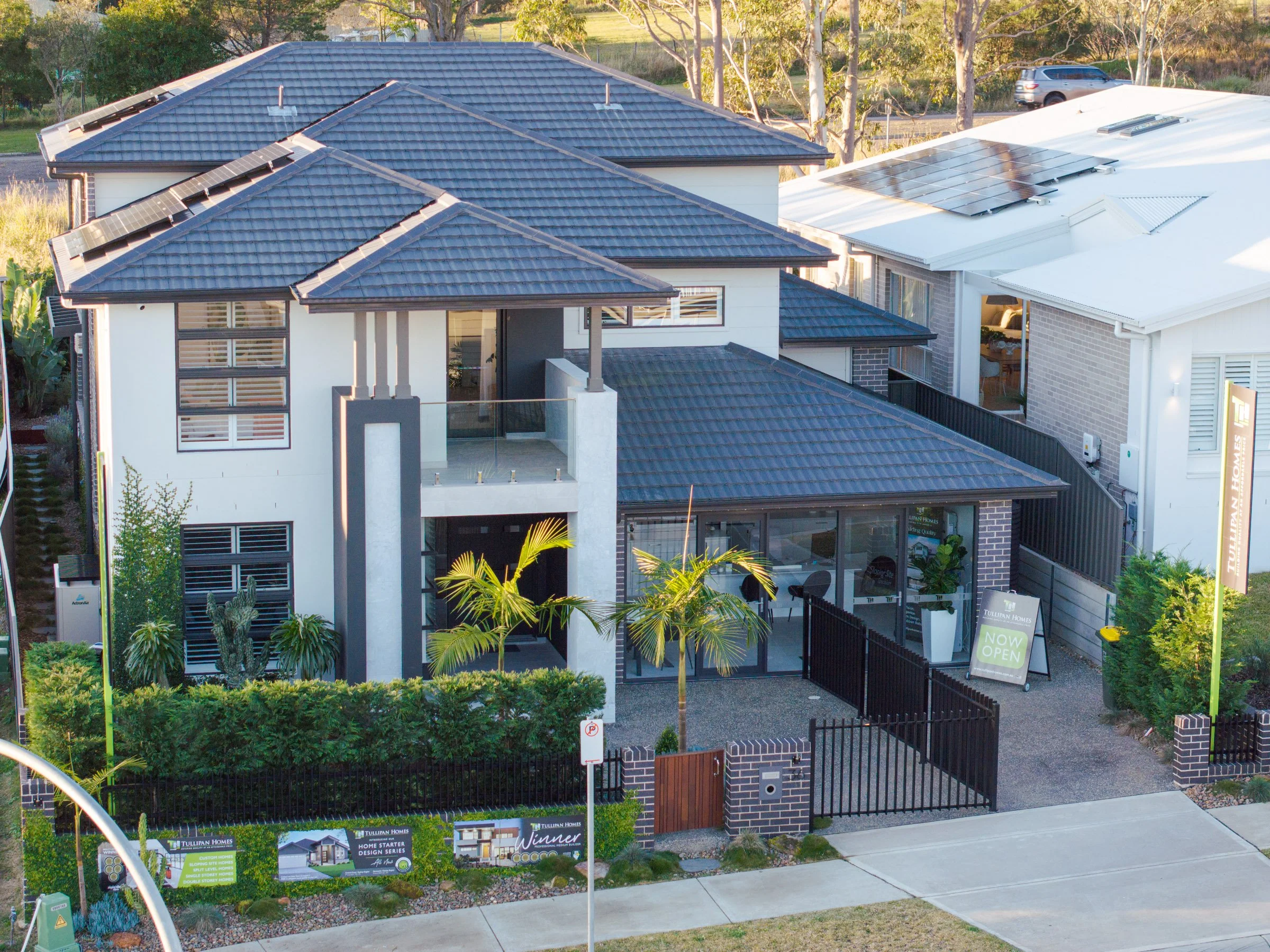 Modern two-story house with a blue tiled roof, large windows, and a front yard with plants, palm trees, and black fencing.