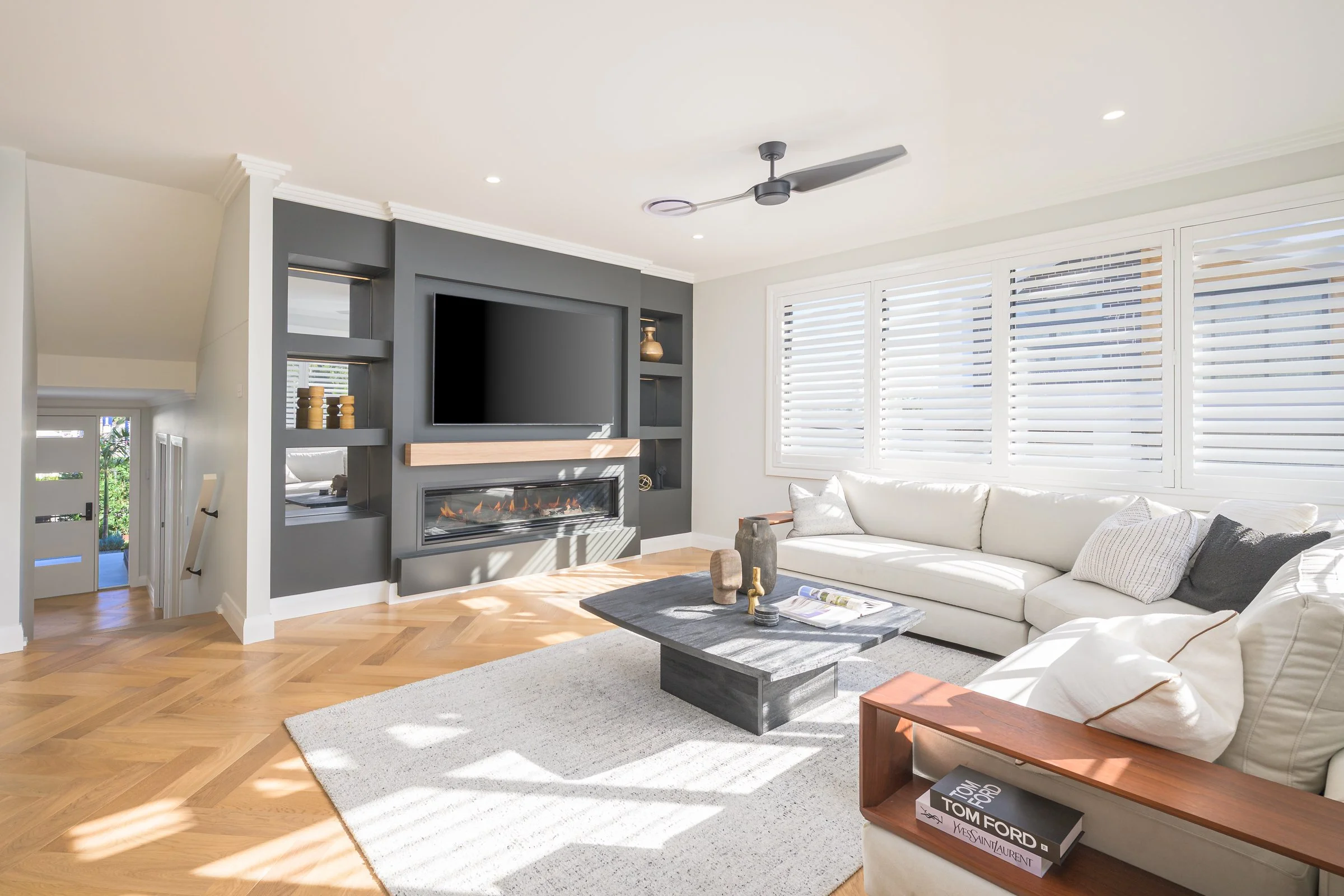 Bright living room with white L-shaped sofa, black coffee table, gray built-in wall unit with TV and fireplace, wood flooring, and large windows with white blinds.