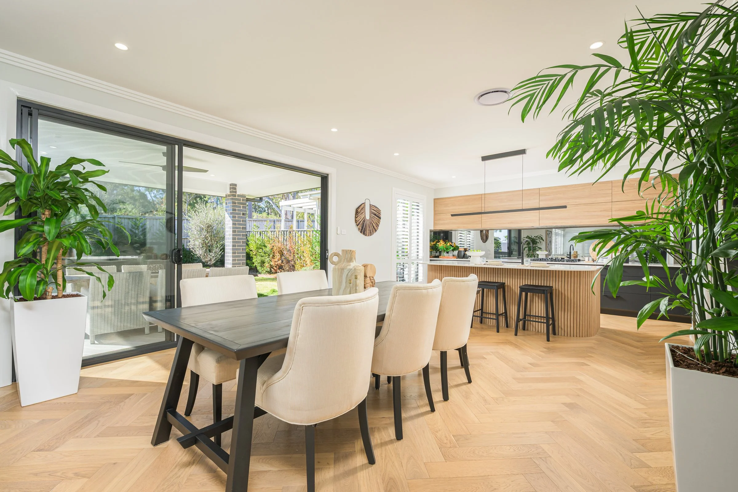 Modern open-concept dining and kitchen area with sliding glass door to garden, wooden flooring, beige upholstered chairs around a dark wooden table, indoor plants, and a kitchen island with bar stools.