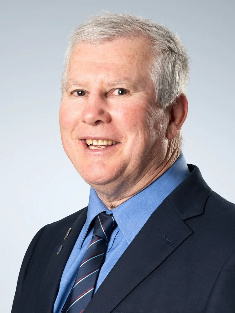 Headshot of a smiling middle-aged man with gray hair, wearing a navy suit, light blue shirt, and striped tie, against a plain light background.