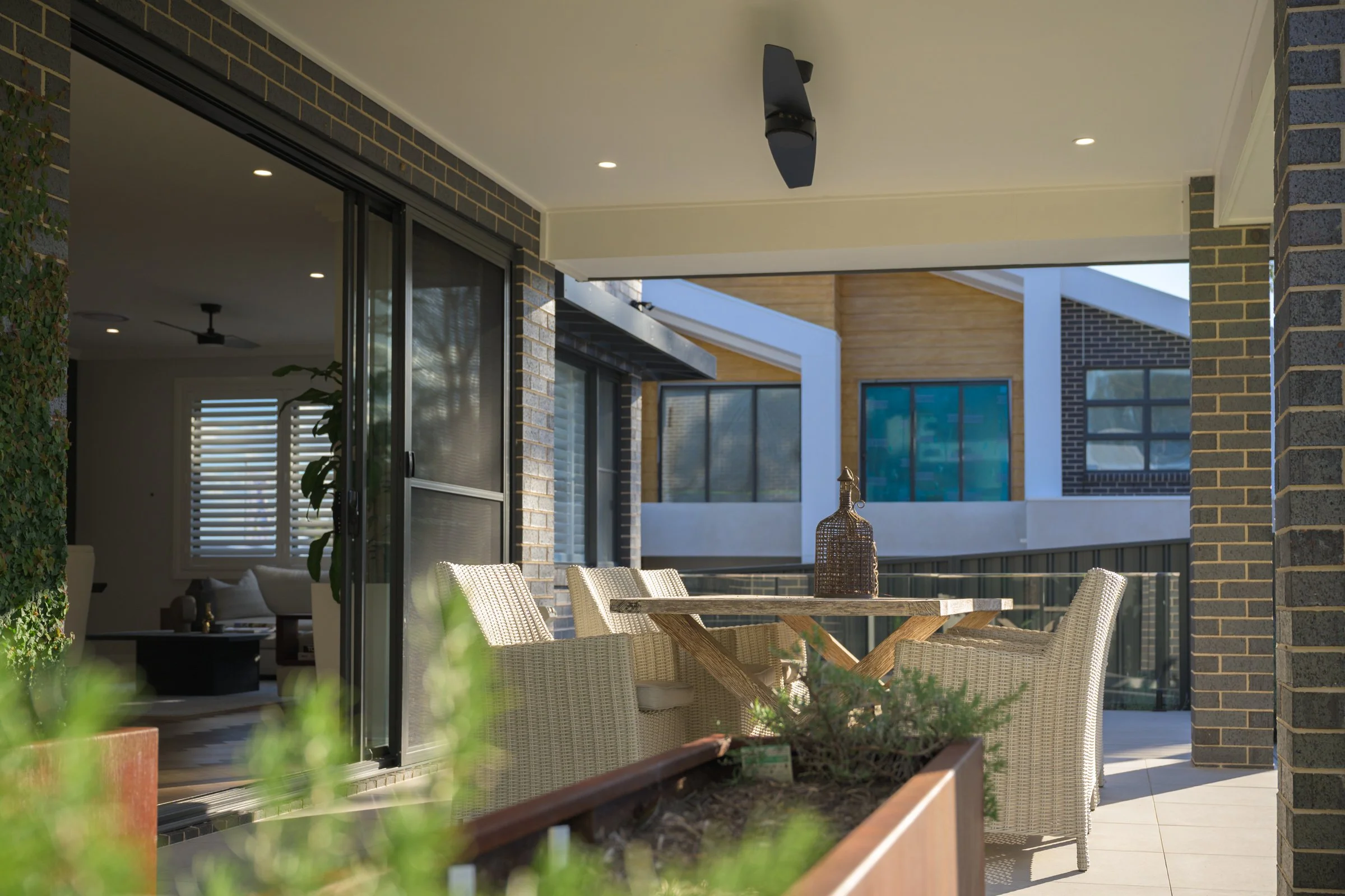 Outdoor patio with wicker chairs around a wooden table and potted plants, adjacent to a modern house with large sliding glass doors and brick walls.
