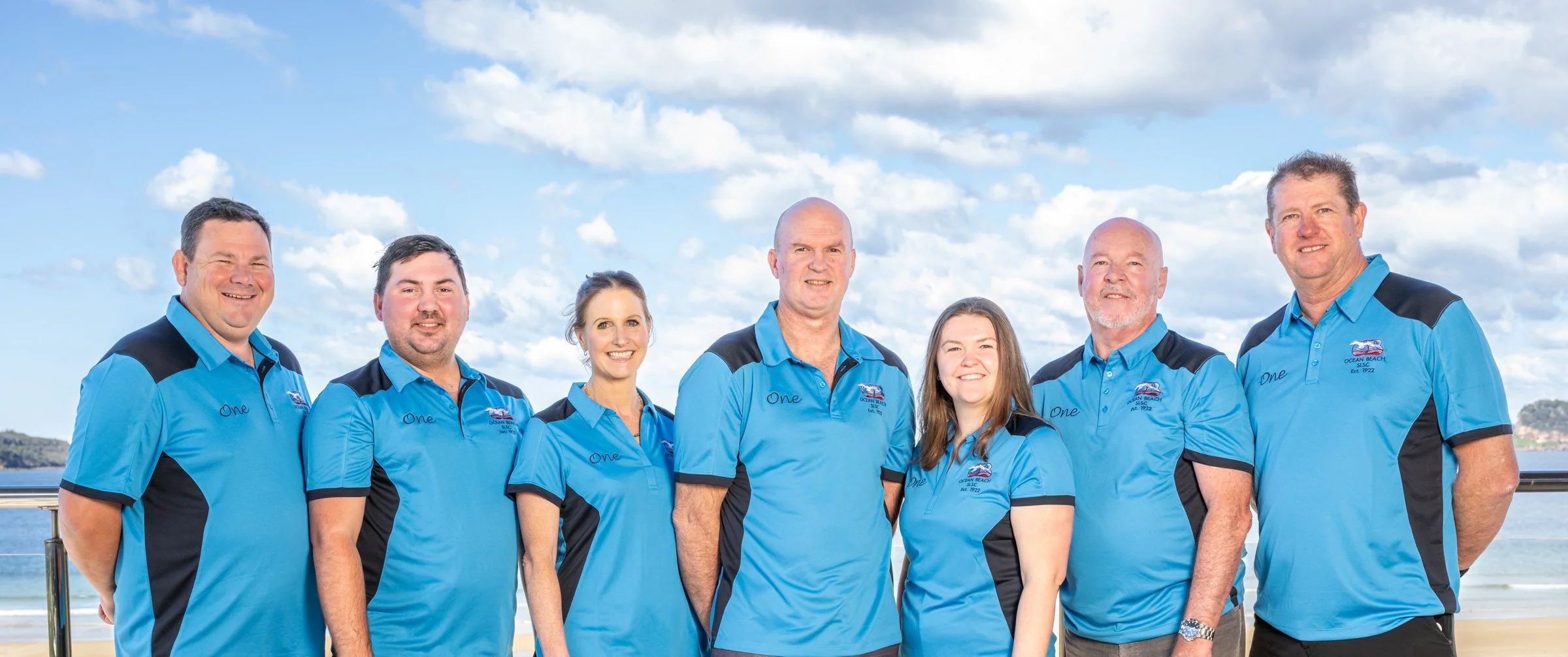 Group of seven people wearing matching blue and black shirts, standing on a beach with ocean and blue sky in the background.