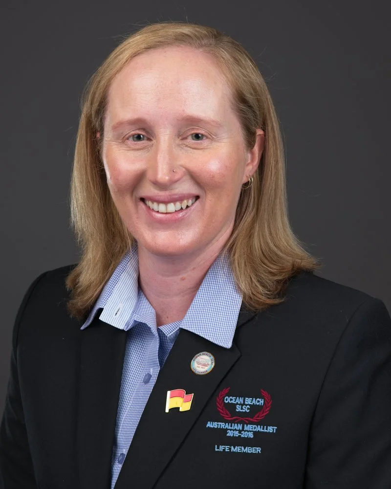 A woman with blonde hair wearing a black blazer with a badge and pins, smiling, against a dark background.