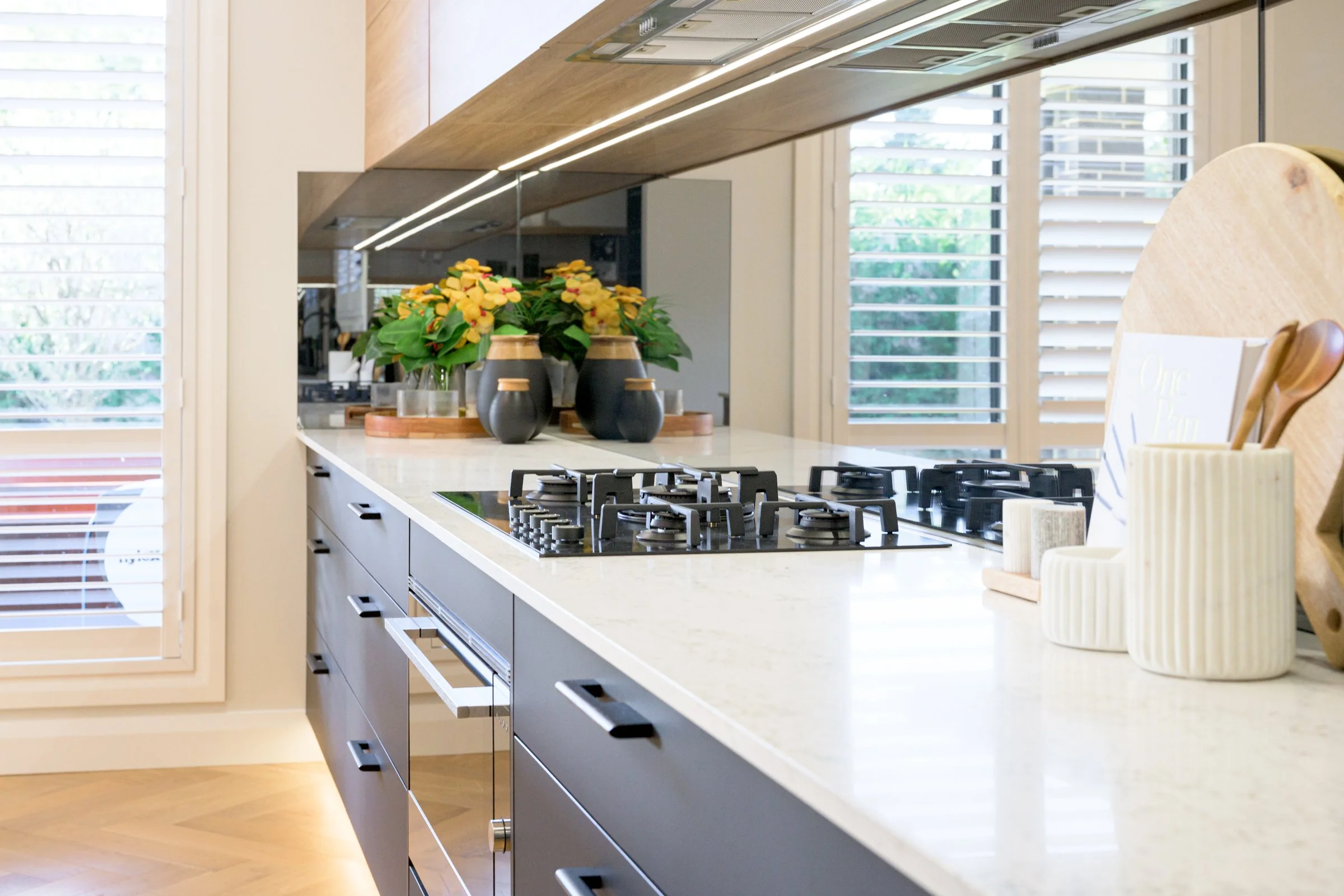 Modern kitchen with white countertop, black kitchen drawers, gas stove, and large window with white blinds. Decorative black vases and yellow flowers on the counter.