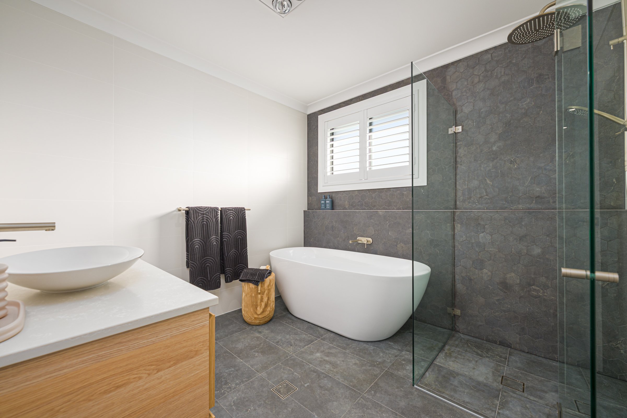 Modern bathroom with white bathtub, glass shower enclosure, wooden vanity with vessel sink, gray tiled floor, and dark tiled accent wall with a window.