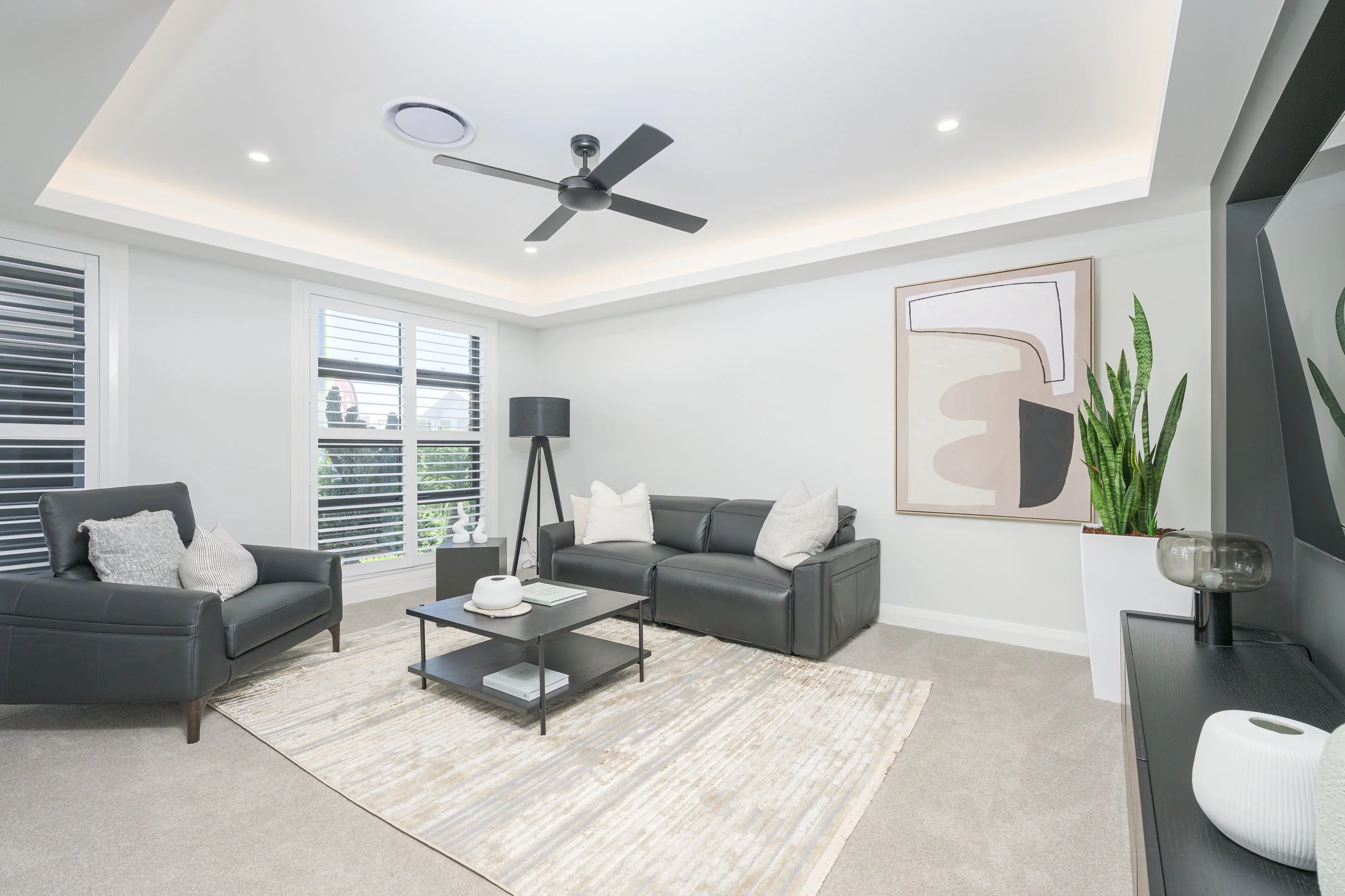 Modern living room with black leather sofas, a black coffee table, a beige rug, artwork on the wall, a tall green plant, and a black floor lamp near windows with white shutters.